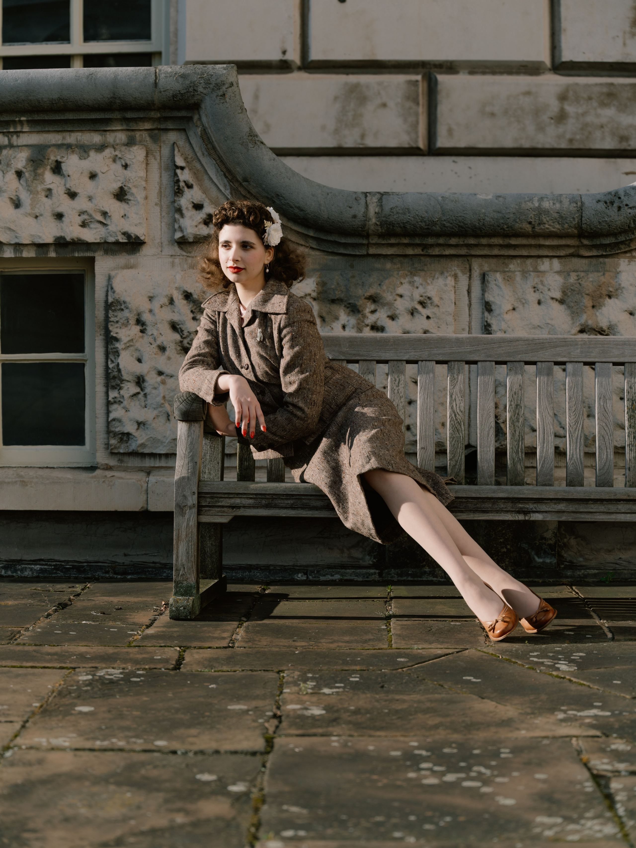 “Wide colour portrait of a woman in a vintage-inspired tweed suit reclining across a wooden bench in warm afternoon sunlight.”
