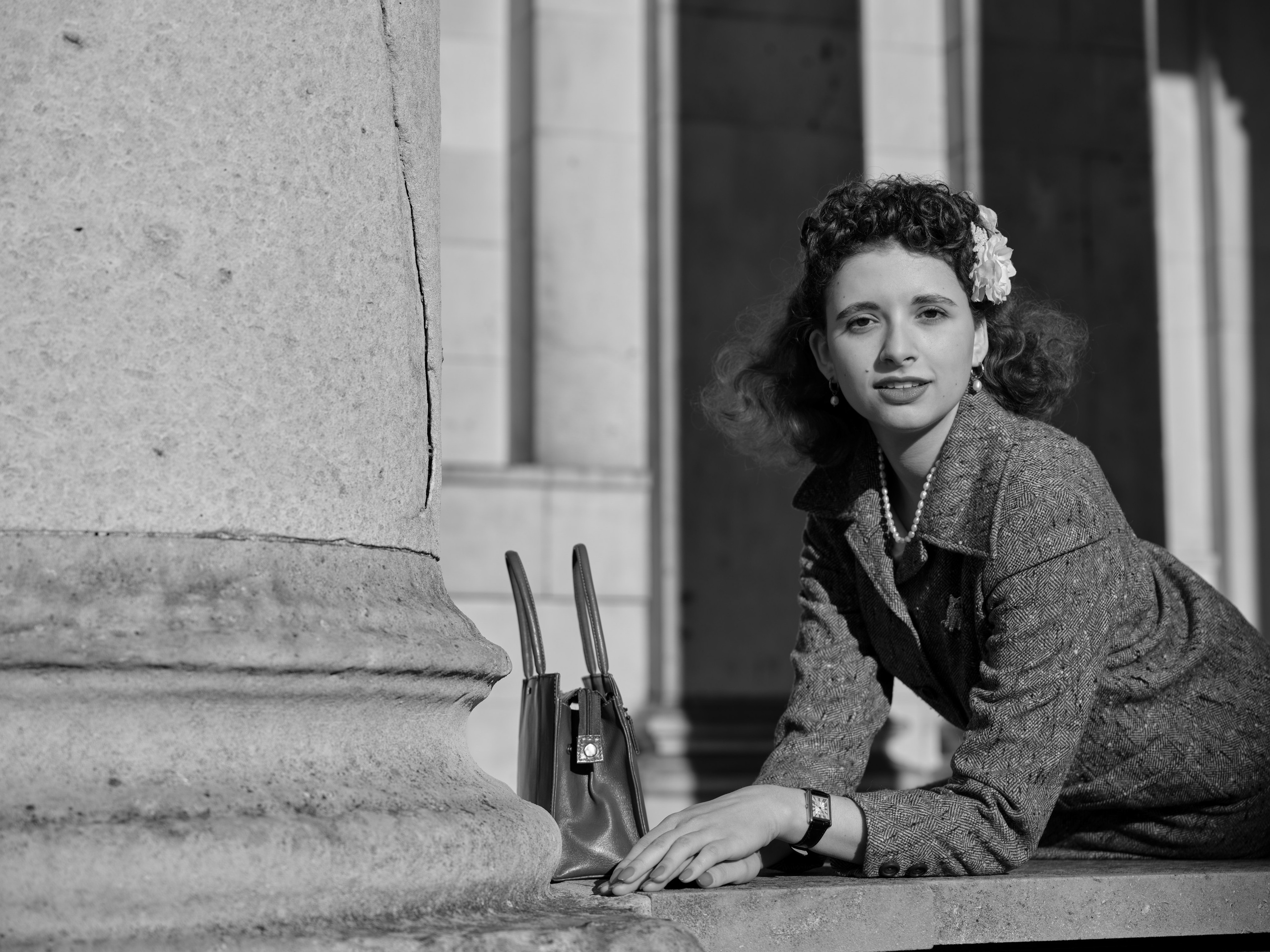 “Black-and-white portrait of a dark-haired woman in a tweed suit leaning on a stone balustrade beside classical architecture.”
