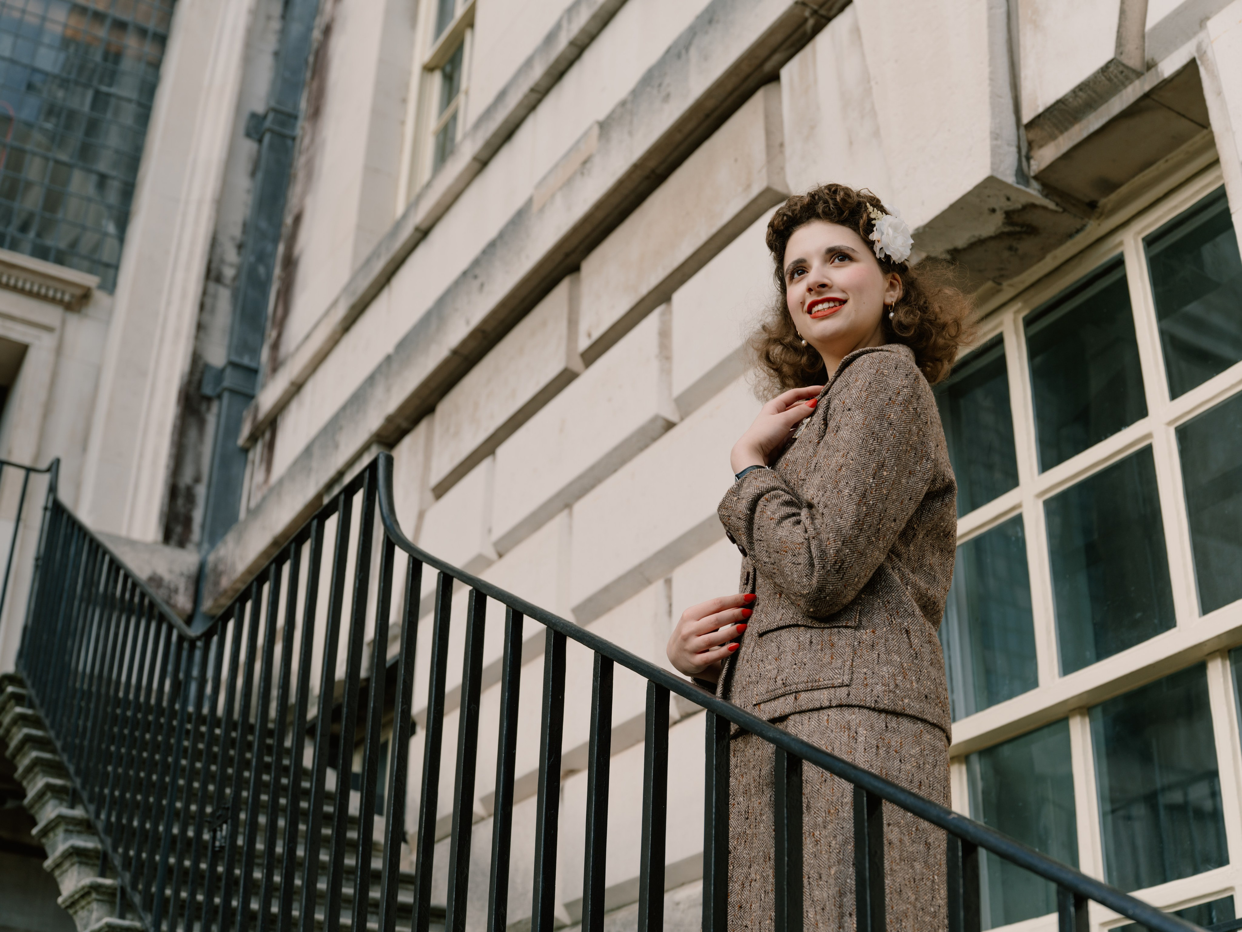 “Low-angle colour portrait of a smiling woman in a brown tweed suit standing beside a black railing and pale stone building.”