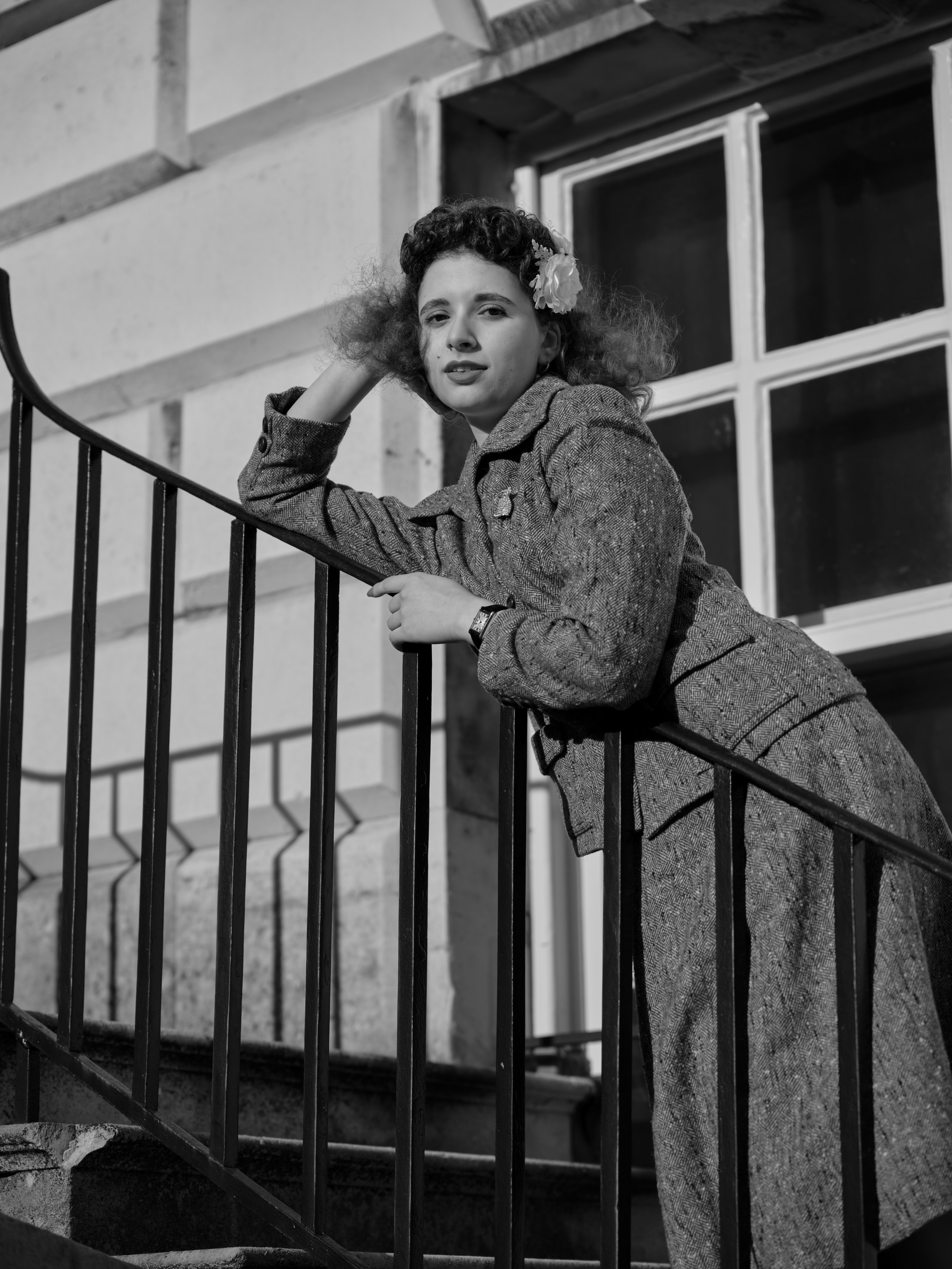 “Black-and-white low-angle portrait of a woman in a vintage-inspired tweed suit leaning against a black metal railing.”