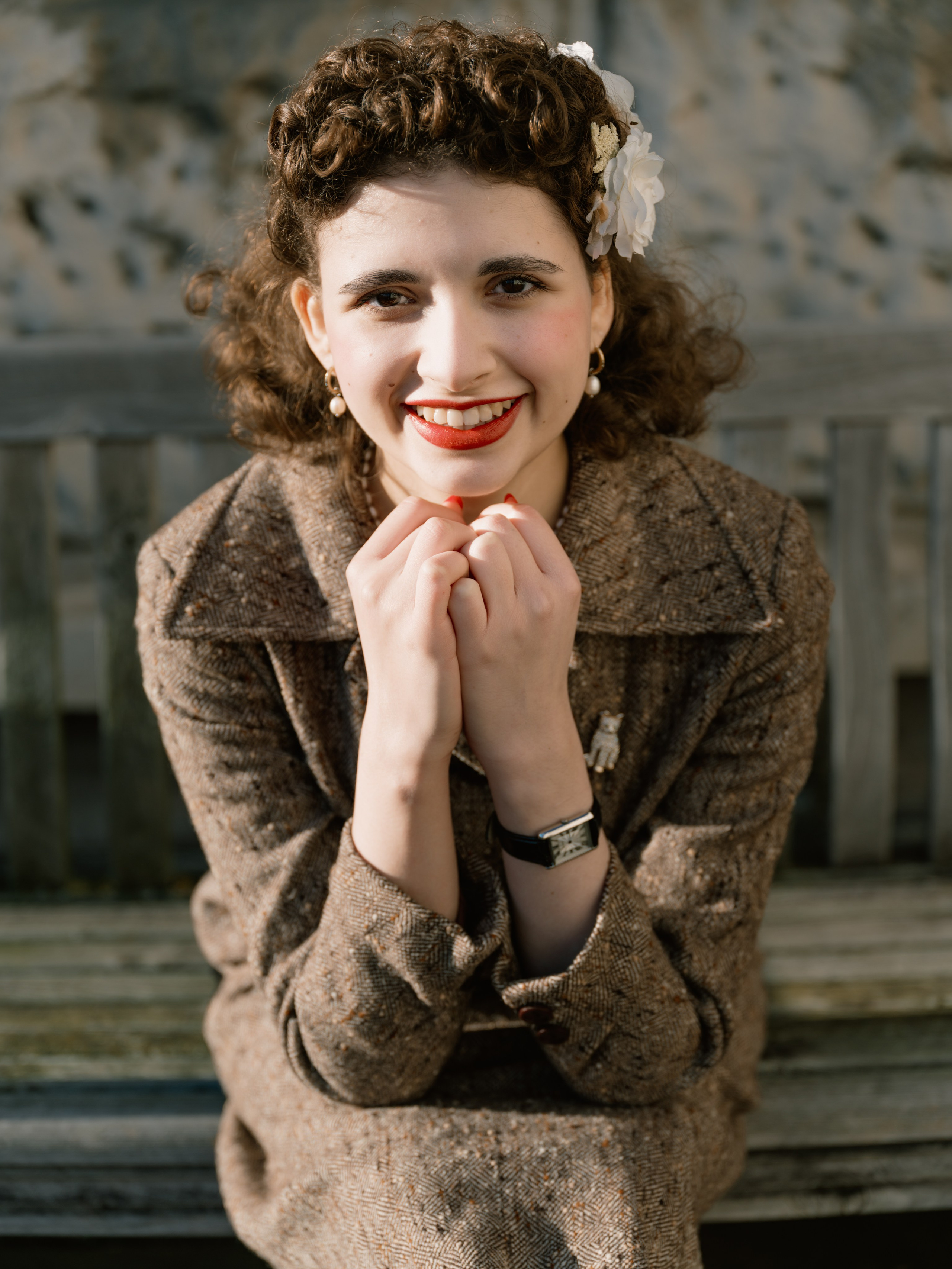 “Wide colour portrait of a woman in a brown vintage-inspired skirt suit sitting along a wooden bench against textured stone architecture.”
