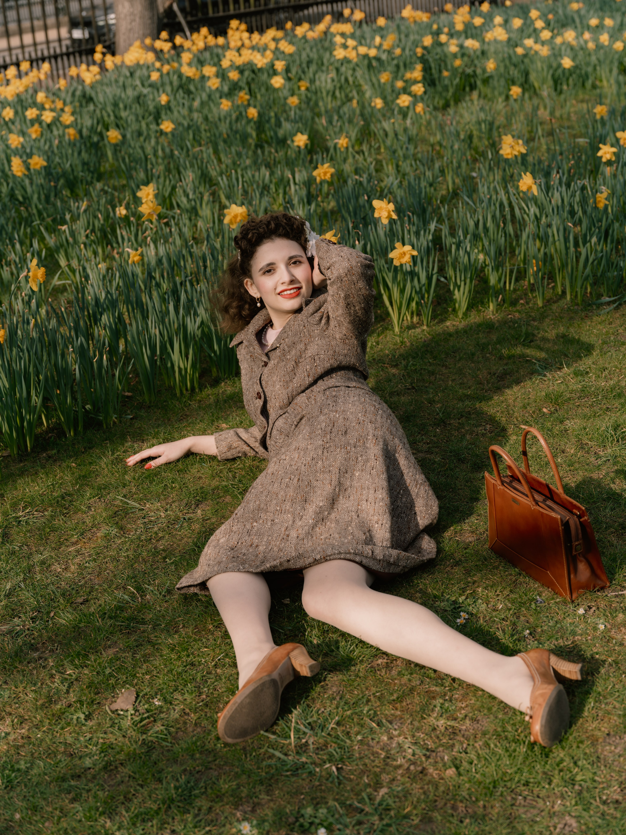“Colour full-length portrait of a woman reclining on the grass beside a brown leather handbag in a garden filled with yellow daffodils.”