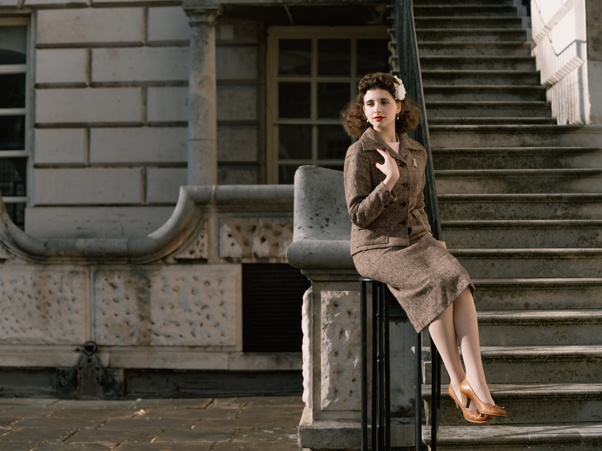 “Full-length colour portrait of a woman in a vintage-inspired brown skirt suit sitting on a black railing beside a broad stone staircase.”