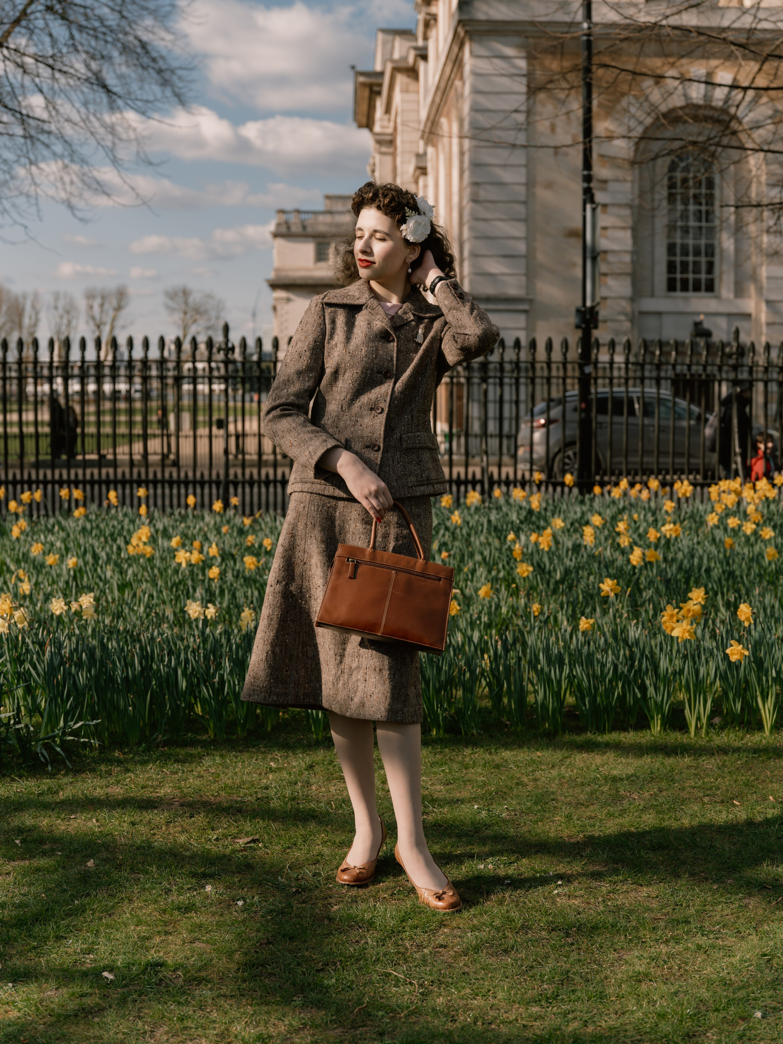 “Full-length colour portrait of a woman in a brown vintage-inspired skirt suit holding a leather handbag among daffodils and historic London architecture.”