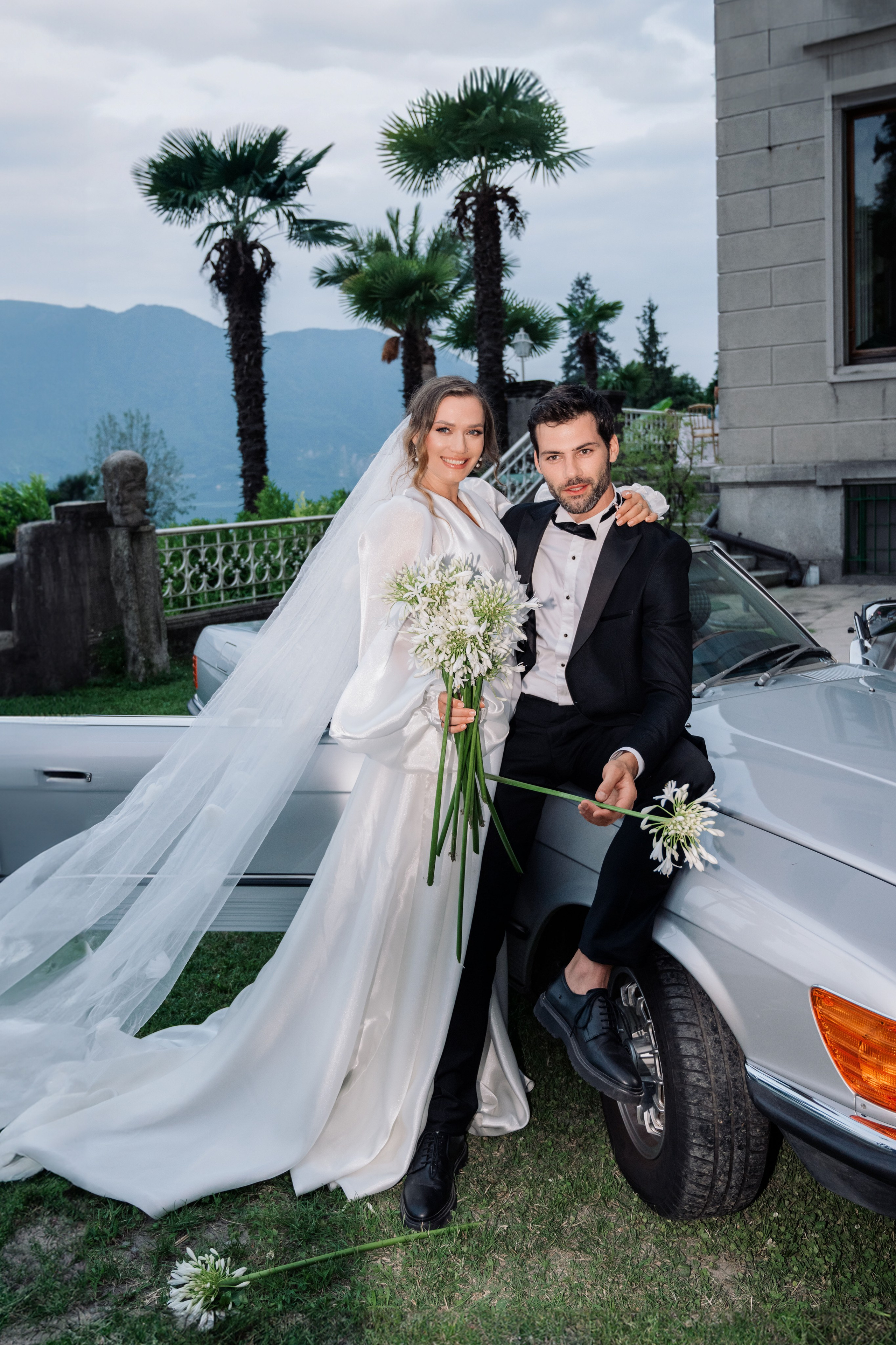 a bride and groom posing in front of a silver car