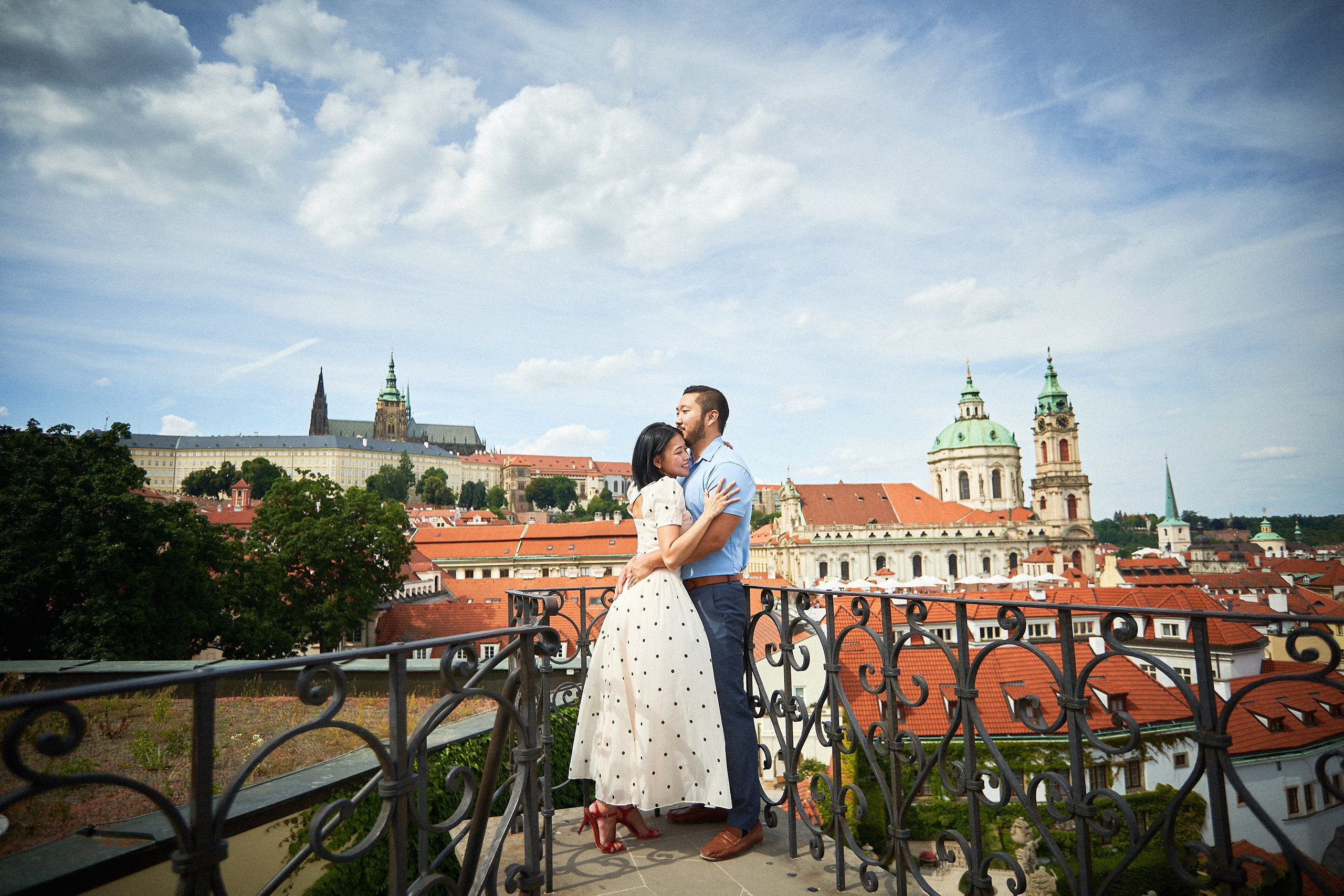 Engaged couple's final embrace against Baroque architecture under a sunny sky in Vrtba Garden.