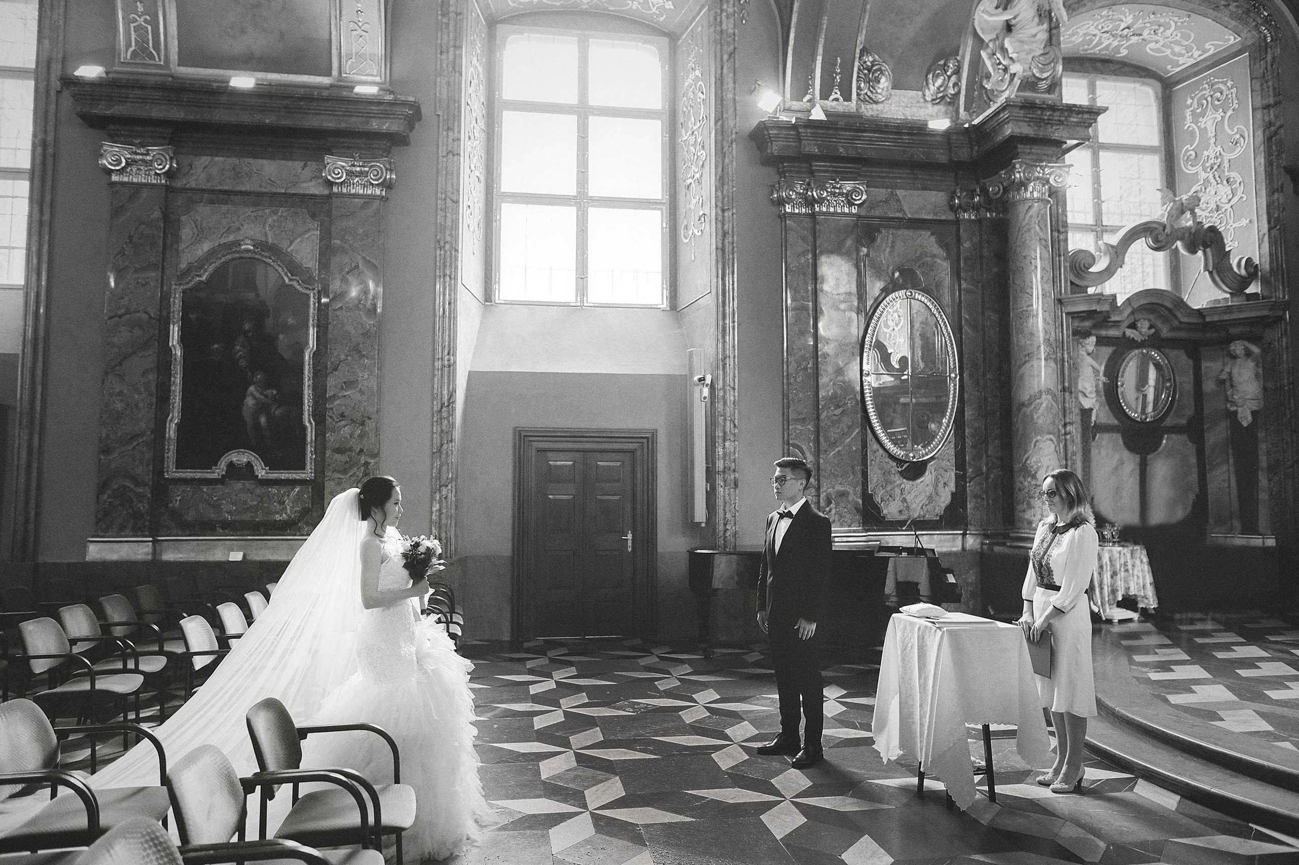 Monochrome image of a bride walking in the soft light from a window at the historic Mirror Chapel in Prague towards her awaiting groom at the altar.