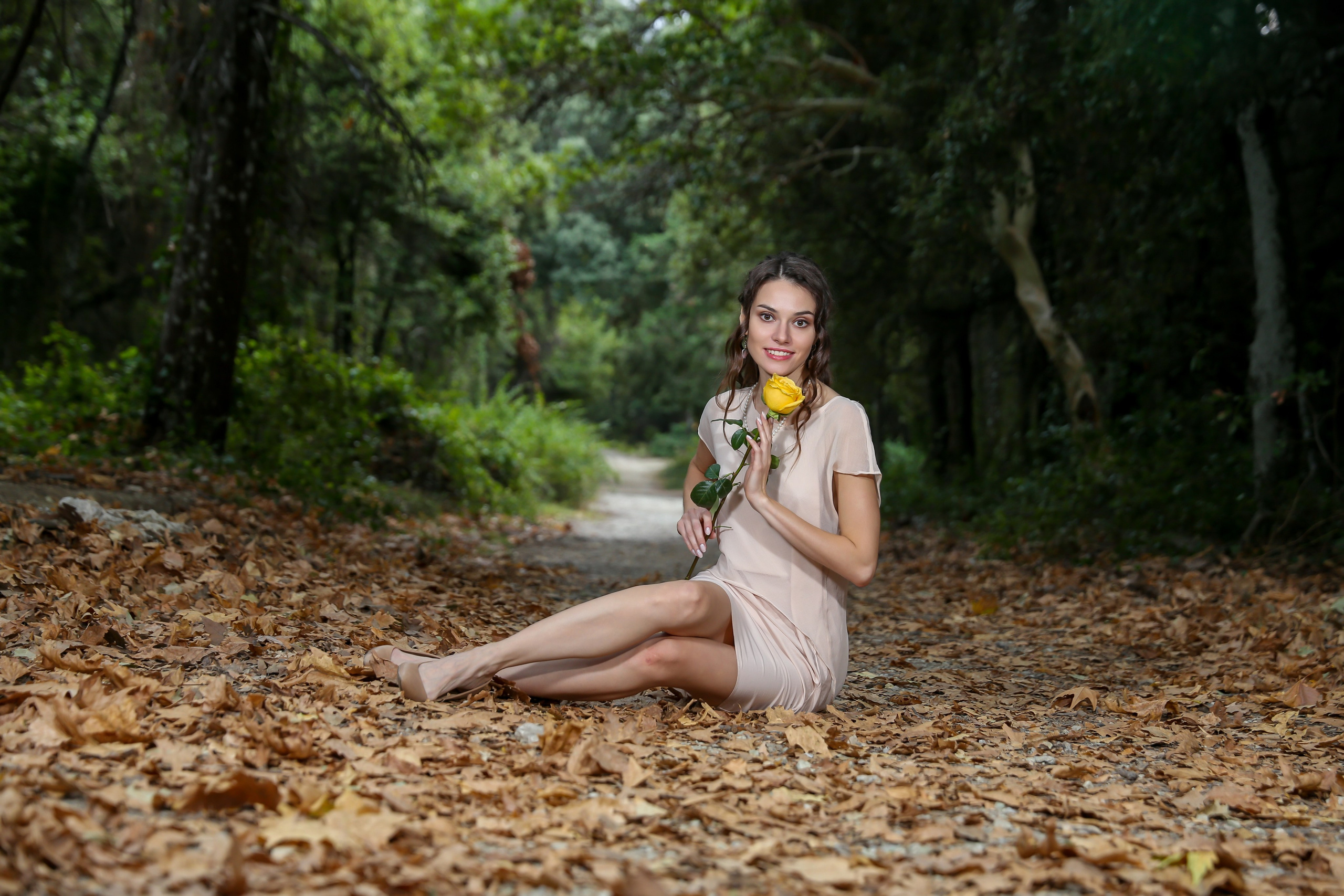 A smiling woman with long brown hair wearing a short beige or light pink dress is sitting on a path covered in dry autumn leaves in a forest, holding a single yellow rose.