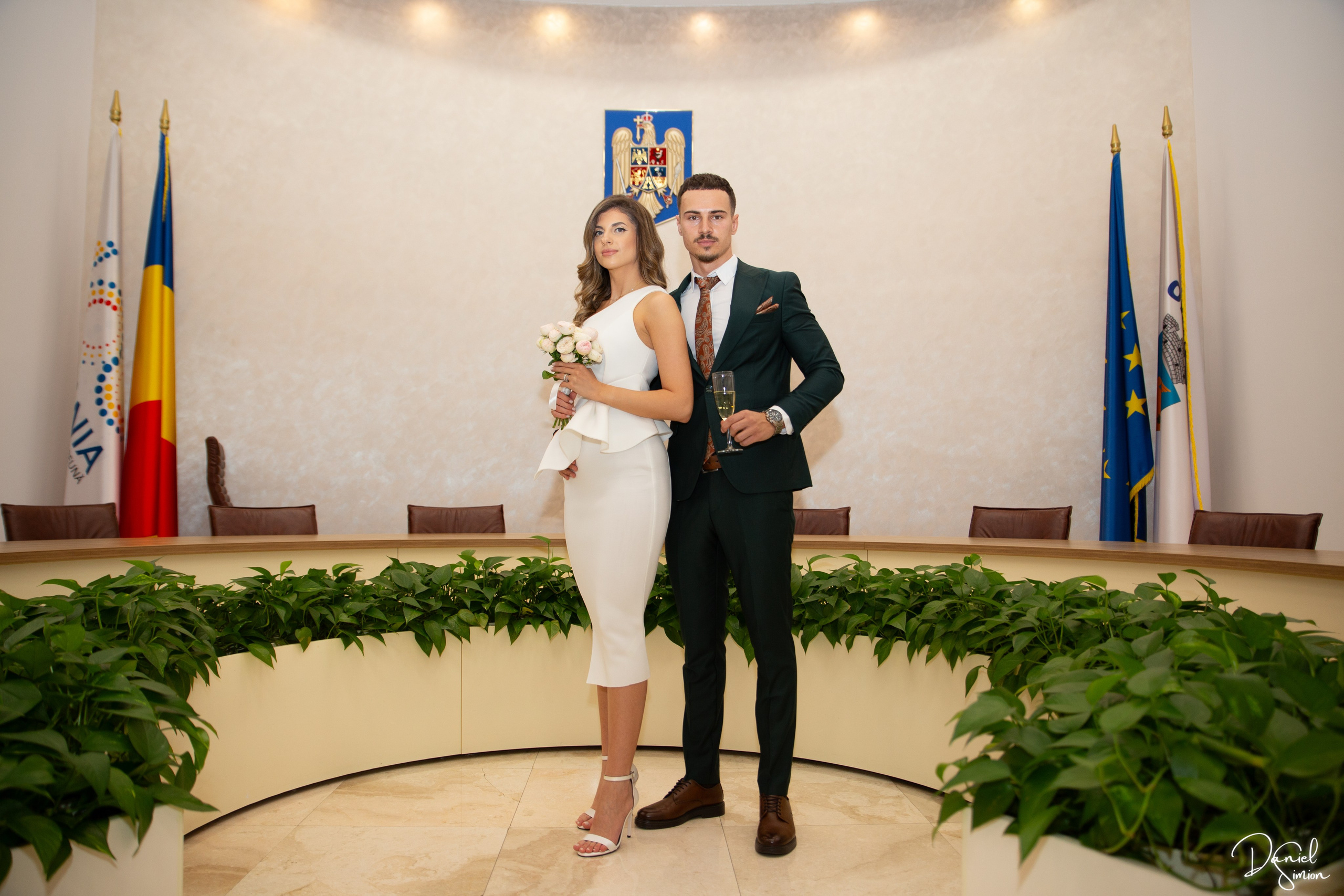 Couple standing indoors in formal attire near flags and official insignia.
