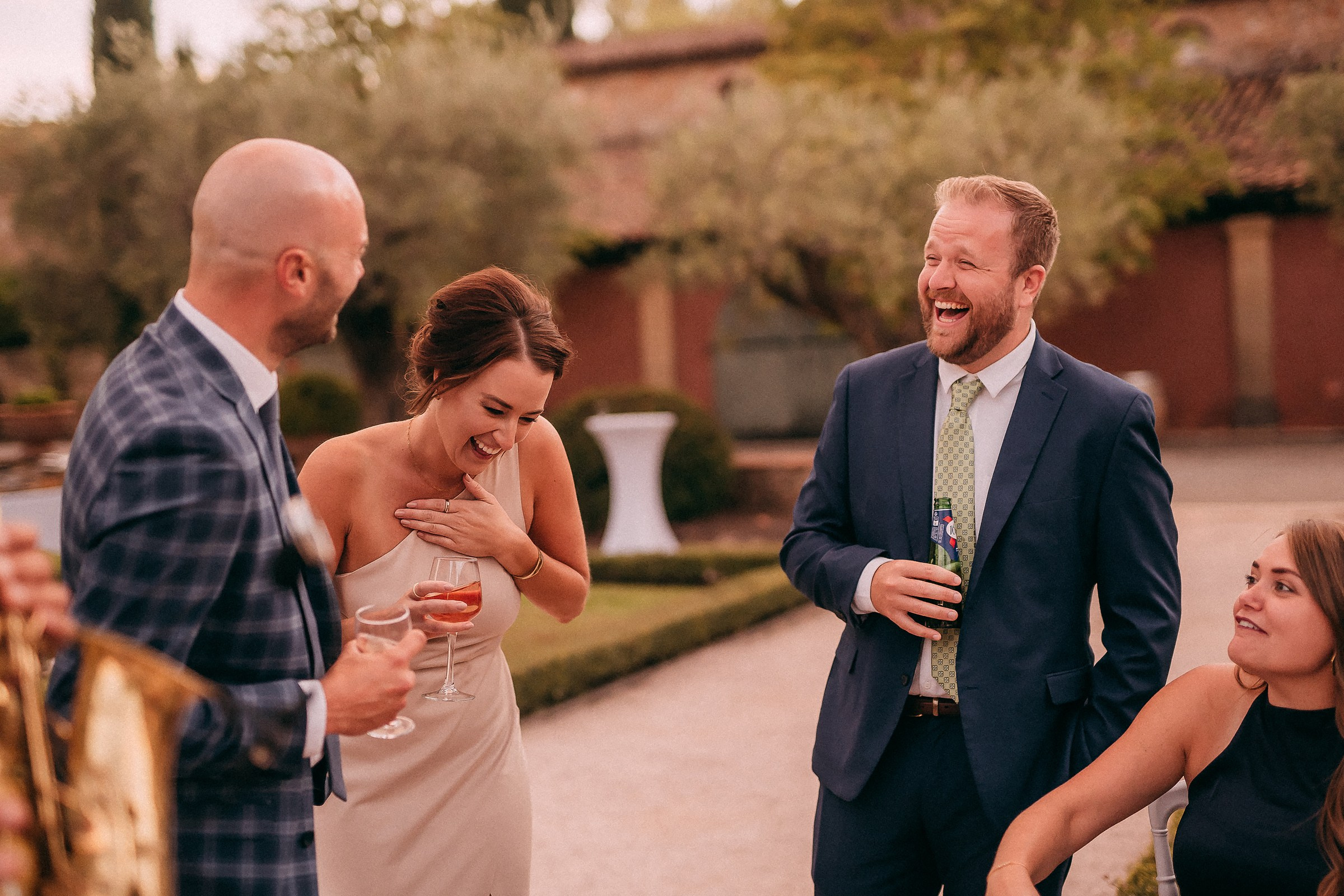 A cheerful group of guests enjoying drinks and laughter during an outdoor wedding celebration, surrounded by manicured gardens. 