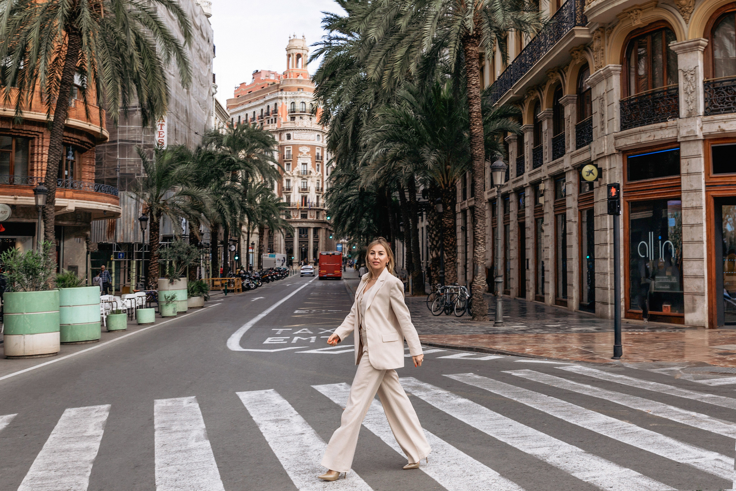 Модная женщина, переходящая пешеходный переход в стильном бежевом костюме, на живописной улице, окруженной пальмами и исторической архитектурой в Валенсии, Испания