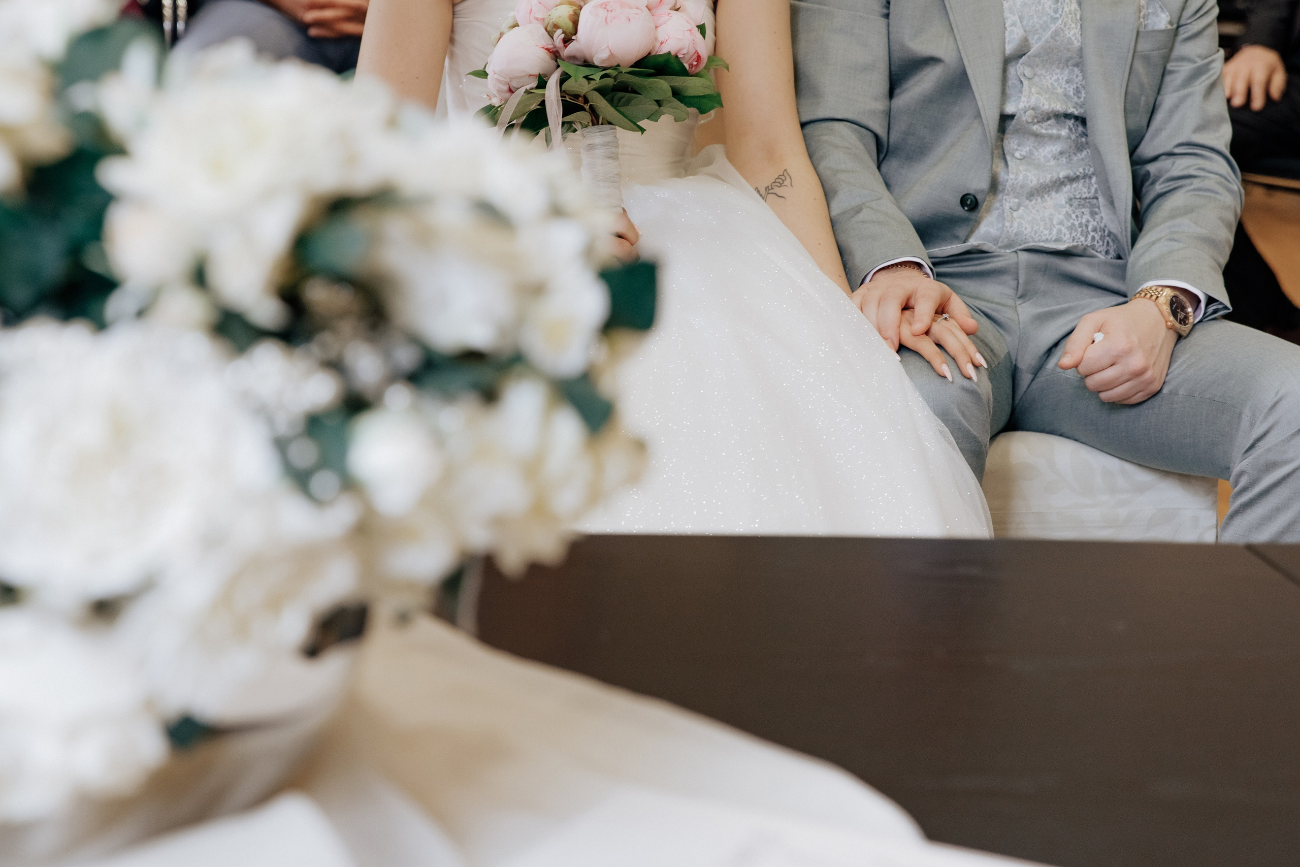 Im Vordergrund ein Brautstrauß, während im Hintergrund die Hände des Bräutigams und der Braut ineinander verschlungen sind.