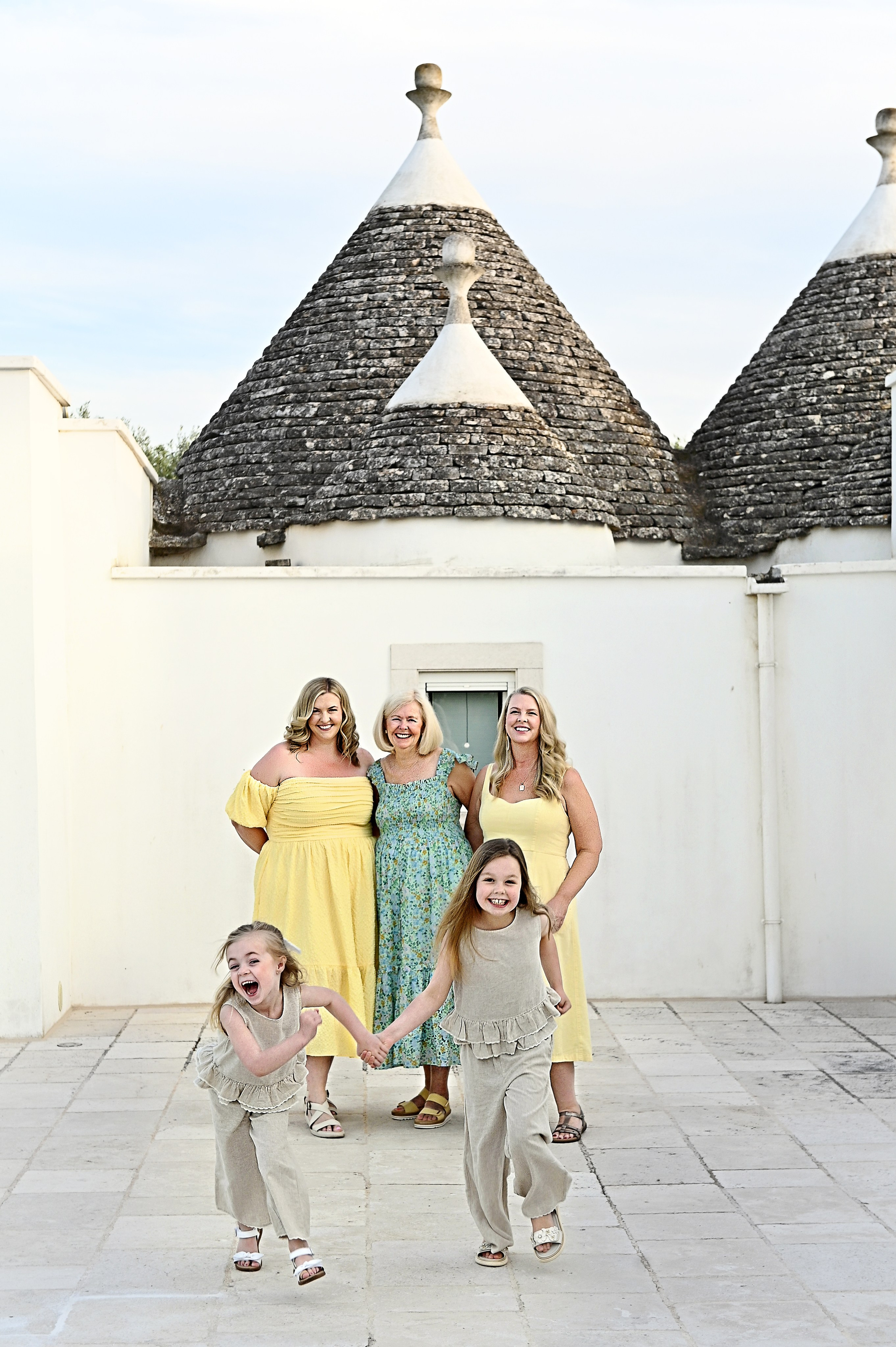 Servizio fotografico di famiglia in Puglia: due bambine felici corrono in avanti mano nella mano, mentre tre donne sorridono sullo sfondo davanti a un tradizionale trullo.