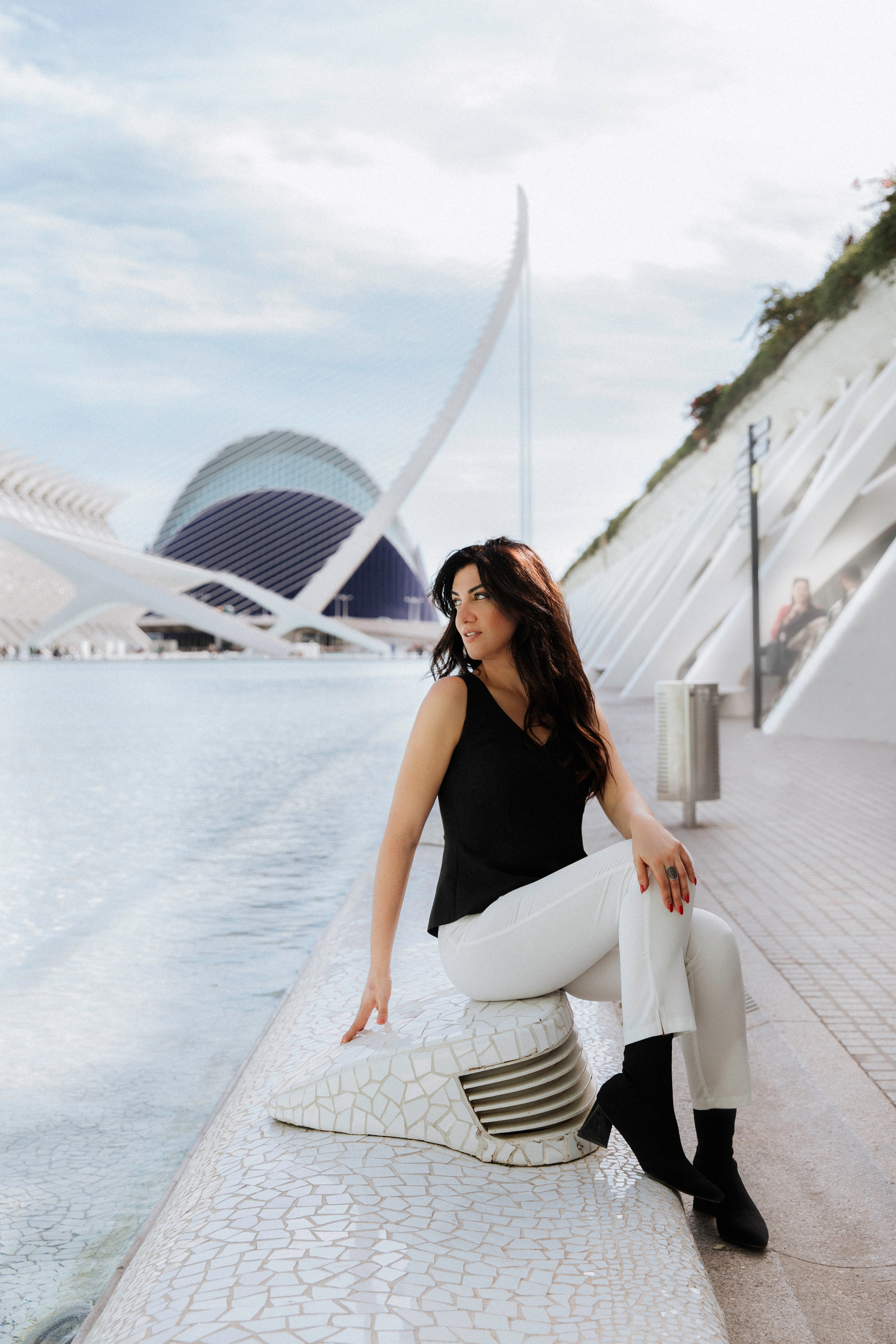 Стильная женщина в черном топе и белых брюках, сидящая у воды в Городе искусств и наук в Валенсии, Испания, на фоне футуристической архитектуры