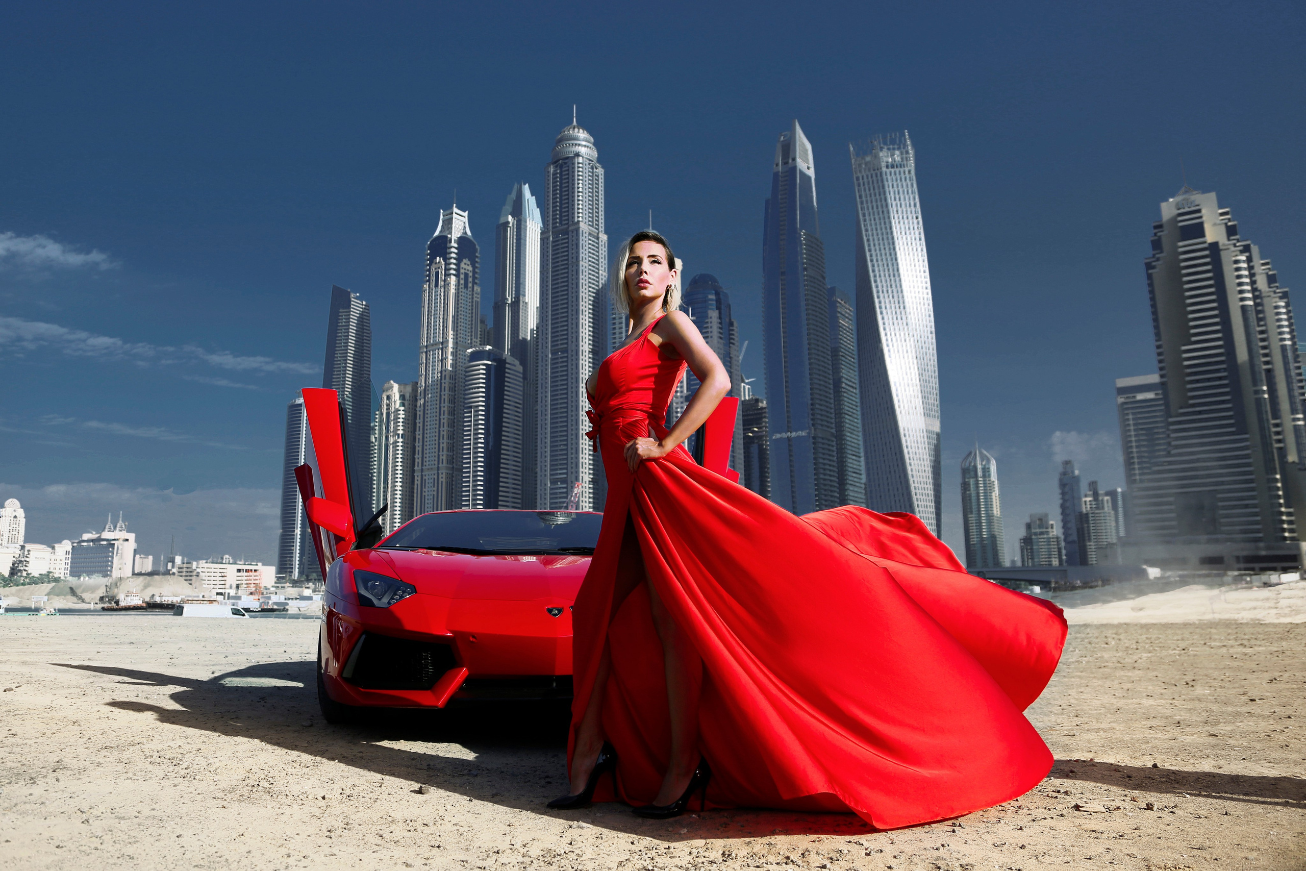 A beautiful model in a red dress stands next to a red Lamborghini with Dubai’s skyscrapers in the background