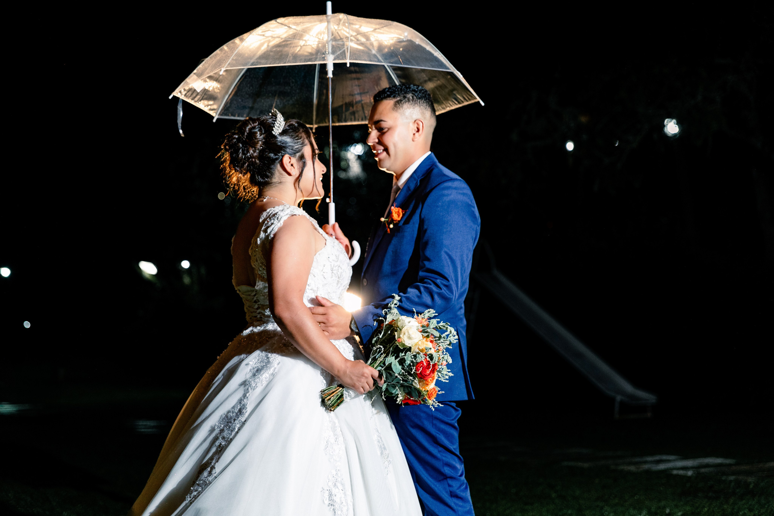 Noiva e noivo sorrindo um para o outro sob guarda-chuva à noite, cena leve e espontânea.
