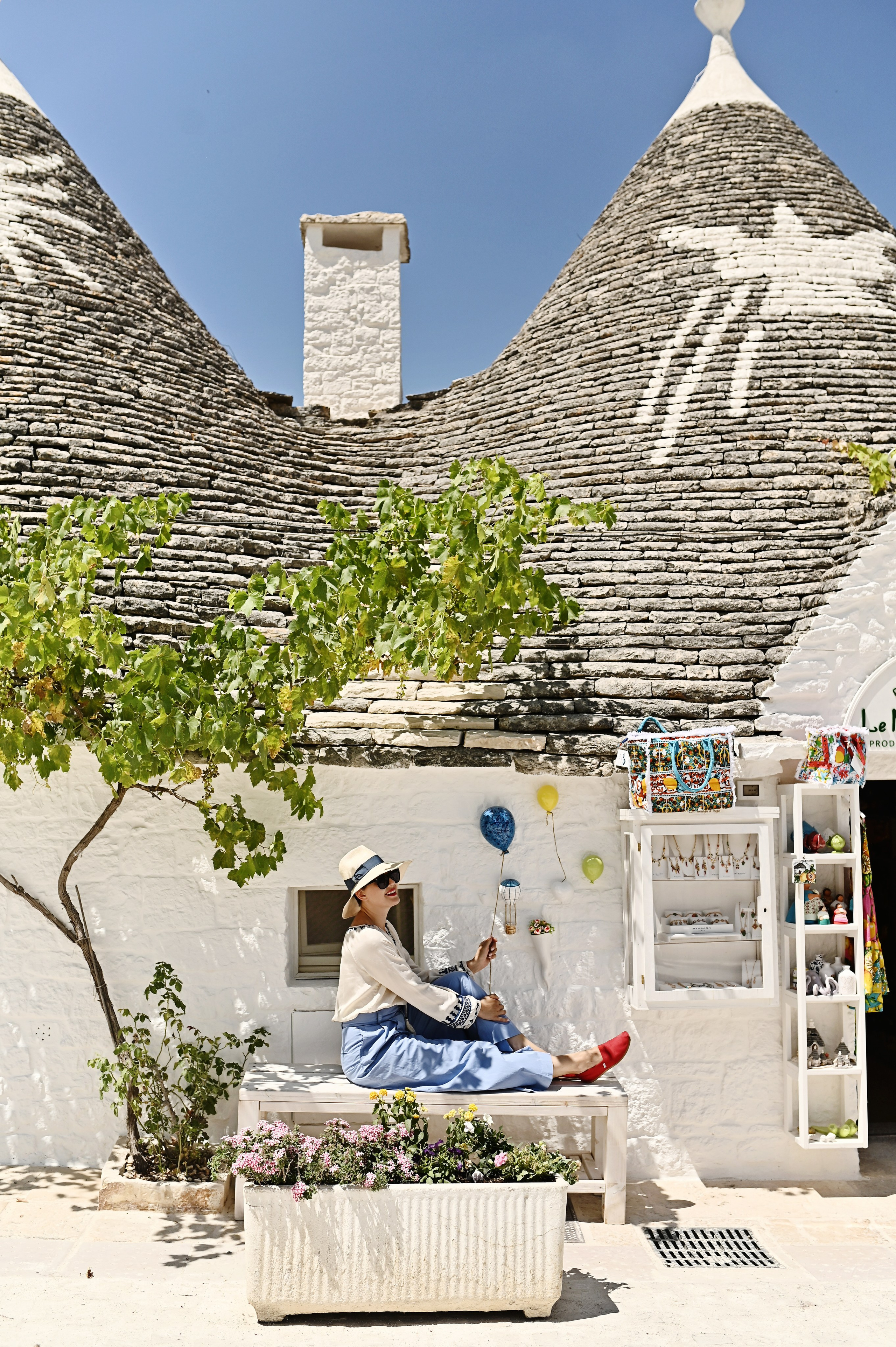 Woman in a straw hat sitting on a bench by a white trullo, holding a decorative balloon.