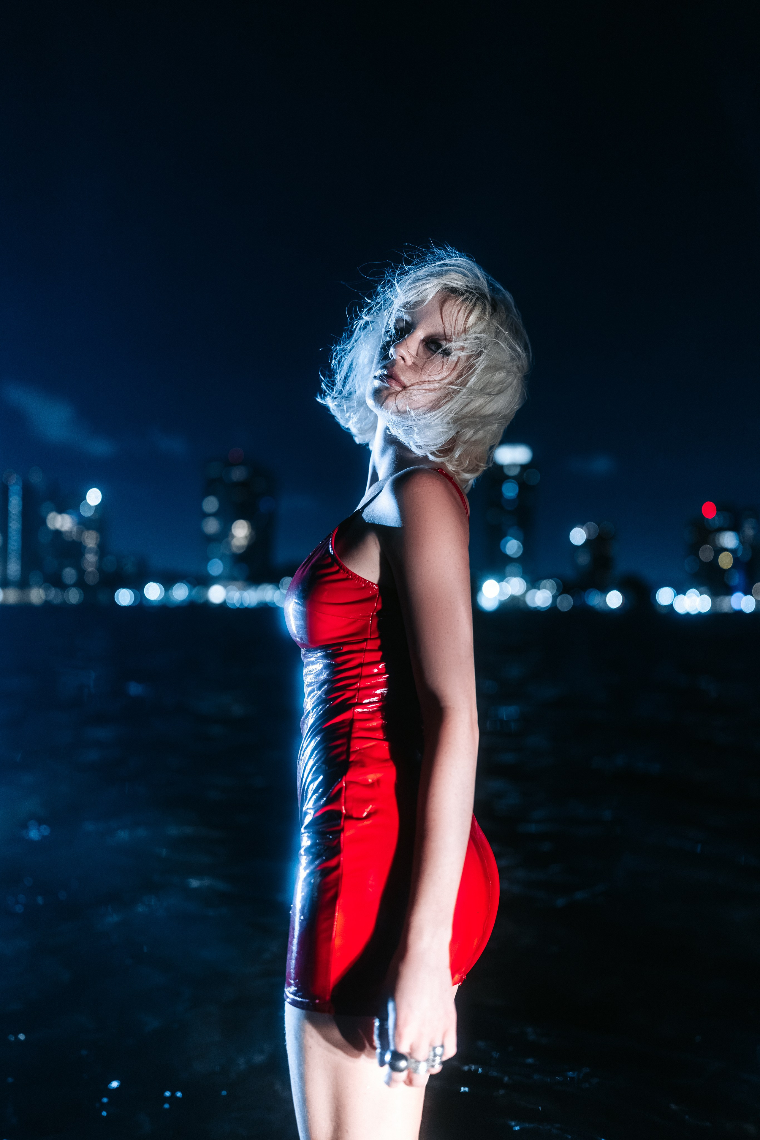 The model stands on the wet shoreline, the red dress lightly clinging to her body. Lighting emphasizes fabric texture and the shine of the water. Confident, cinematic presence.