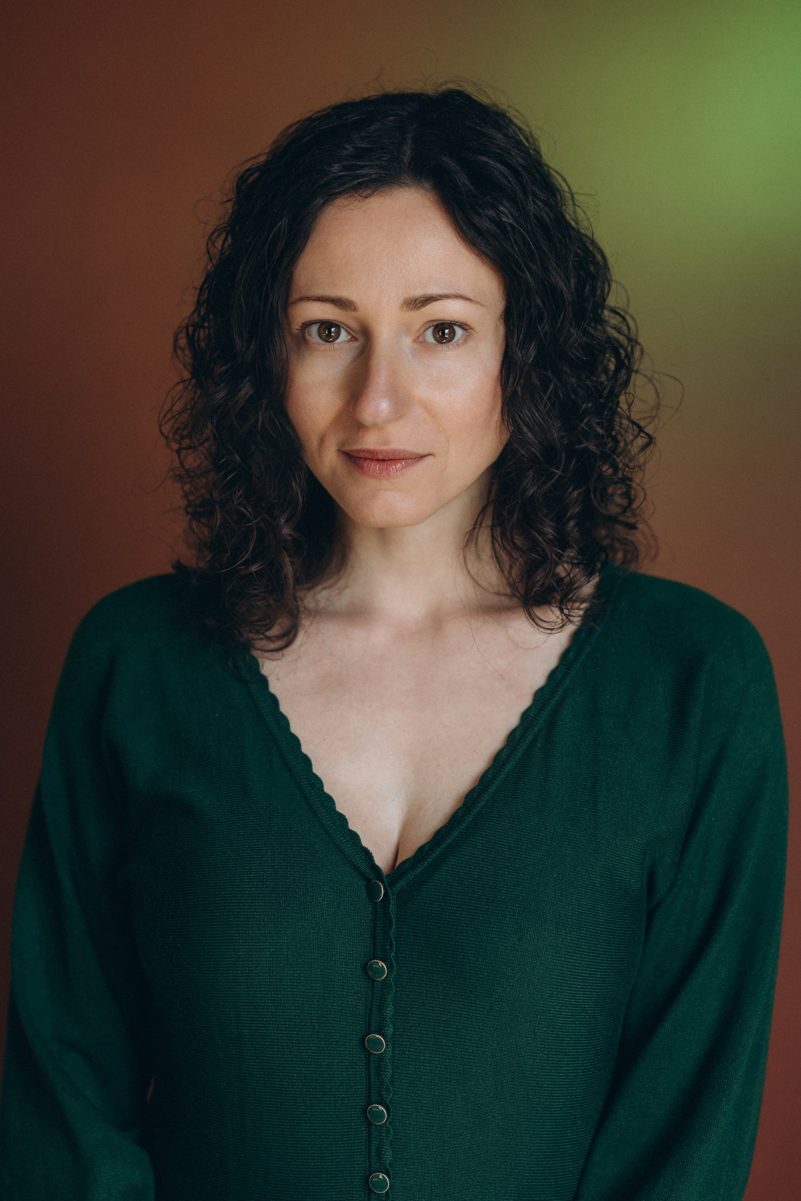 Actor headshot of a woman with curly hair in a green top, turned sideways and looking over her shoulder against a brown background