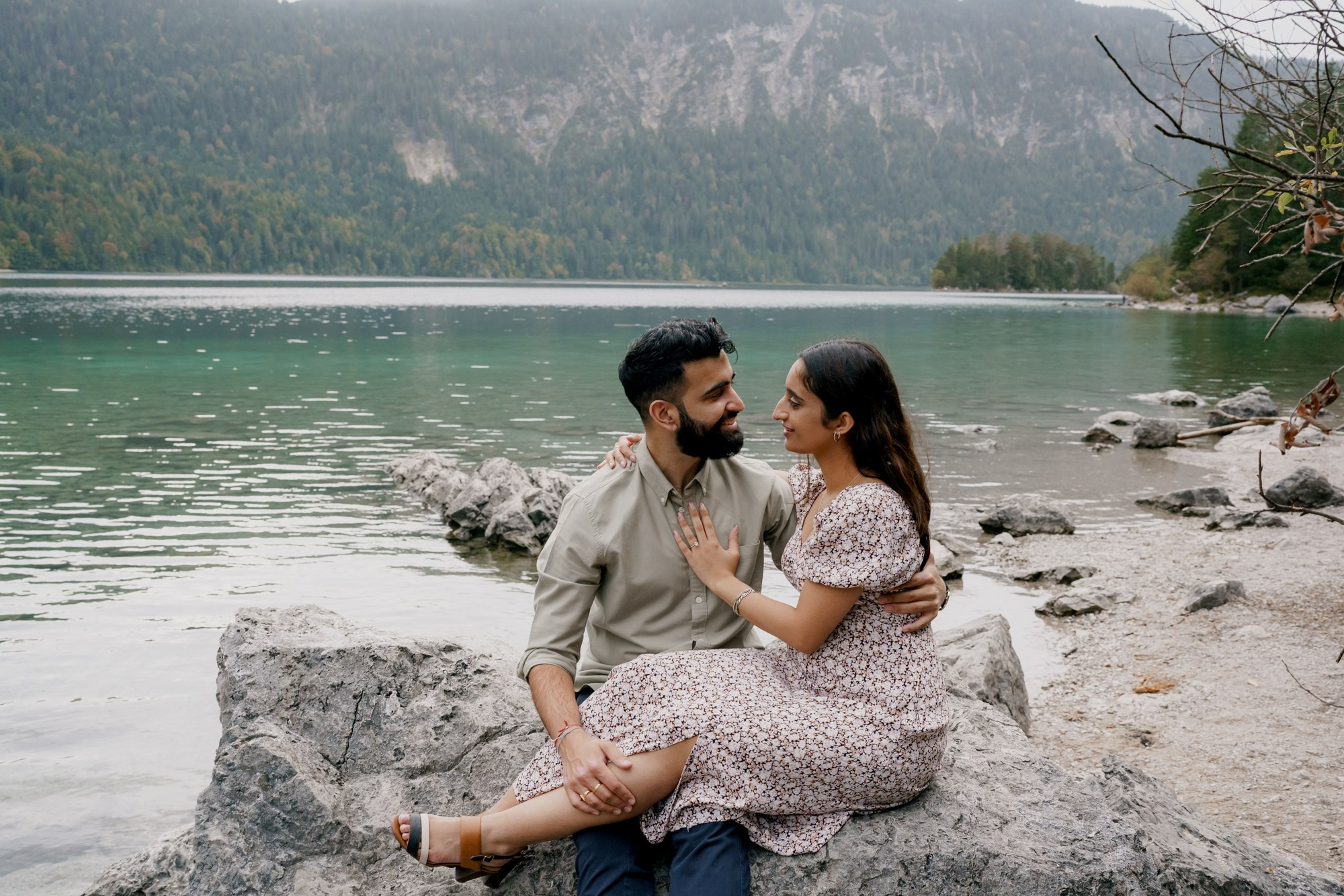 Indian couple are sitting together and looking to each other with mountains and green water of Eibsee at the background 