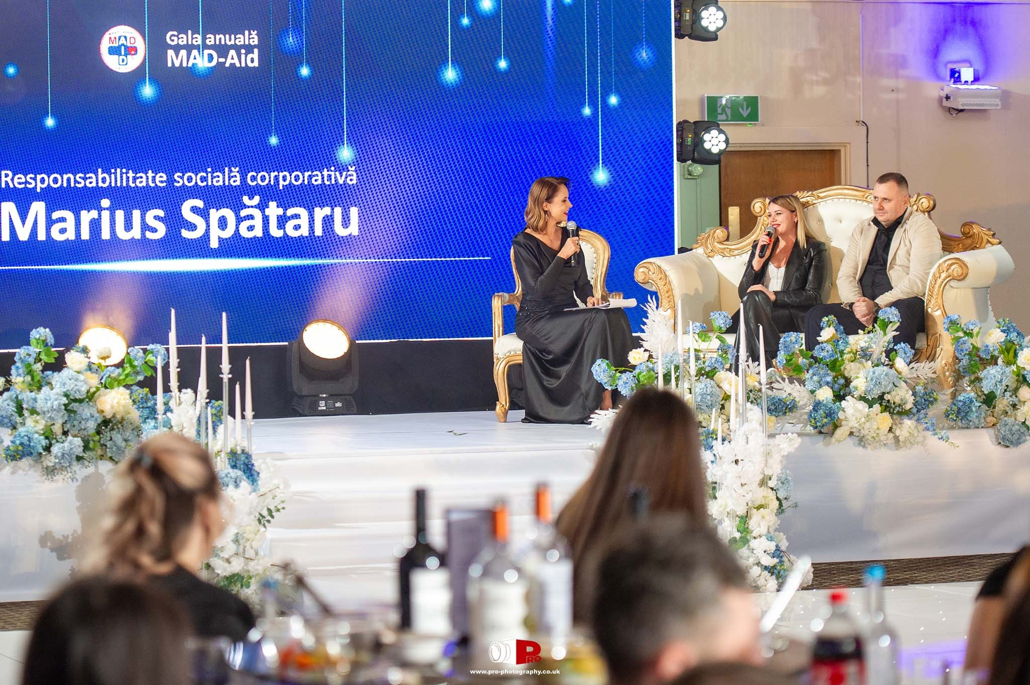 Panel discussion at the MAD-Aid Gala event with a host interviewing two guests on a luxurious stage decorated with blue and white flowers.