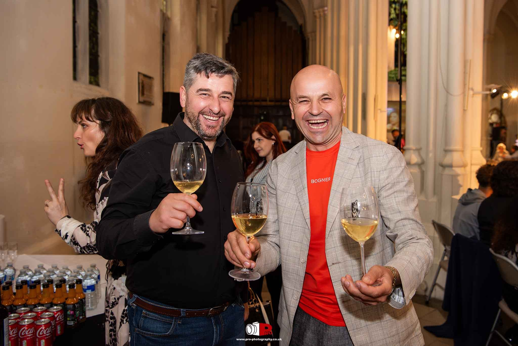 Two smiling men holding wine glasses and celebrating at a lively social gathering.
