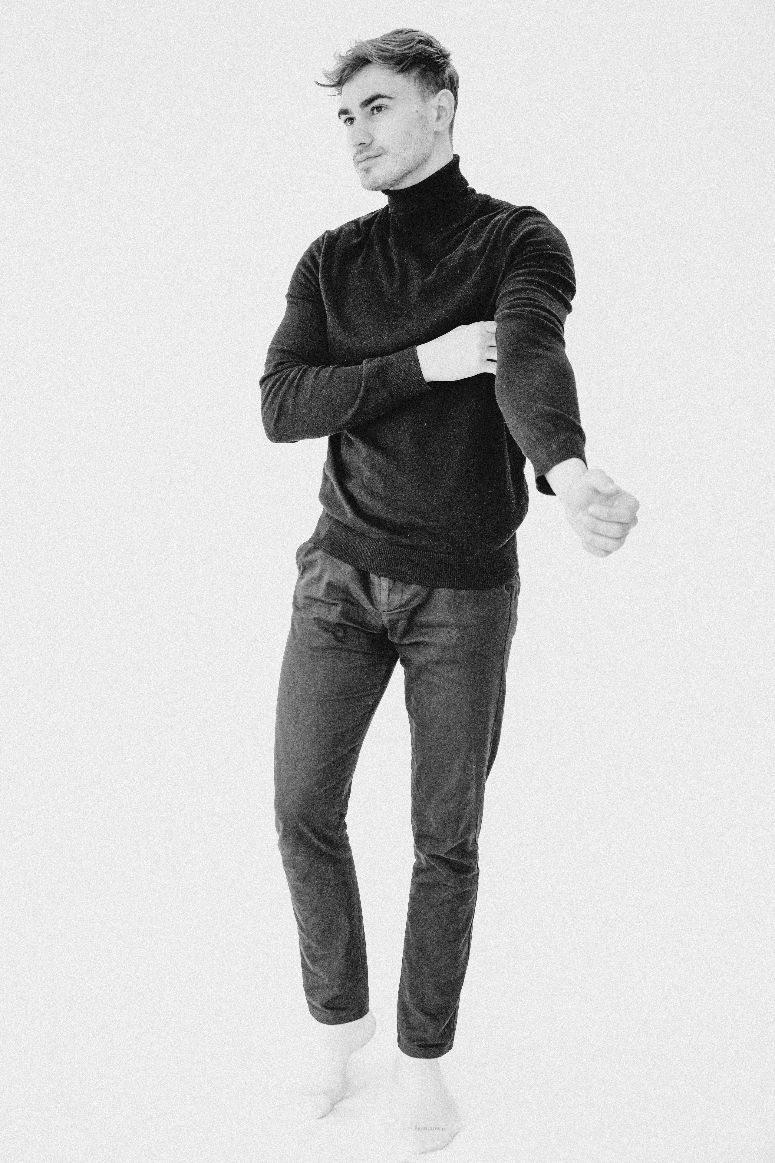 Full-body black and white portrait of a male model standing in a black turtleneck and dark trousers, hand in pocket, against a plain light background.