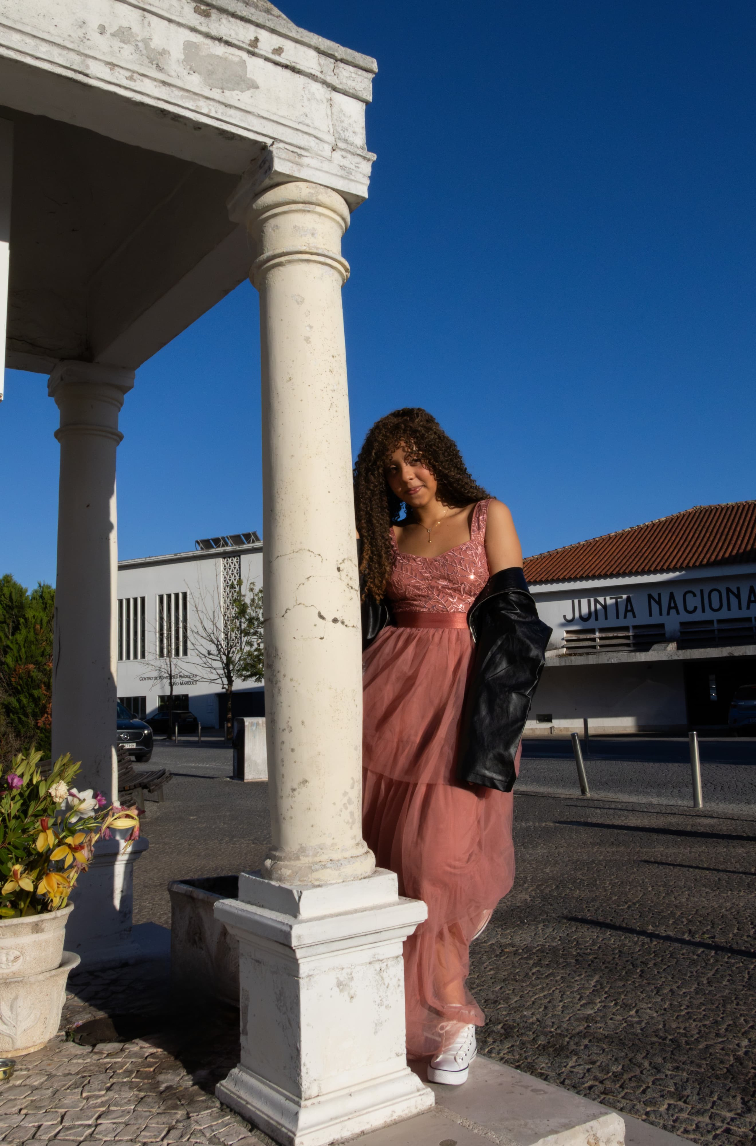 Adolescente com vestido longo rosa caminhando entre colunas brancas de uma estrutura clássica ao ar livre, sob um céu azul limpo. Fotografia de moda conceitual.