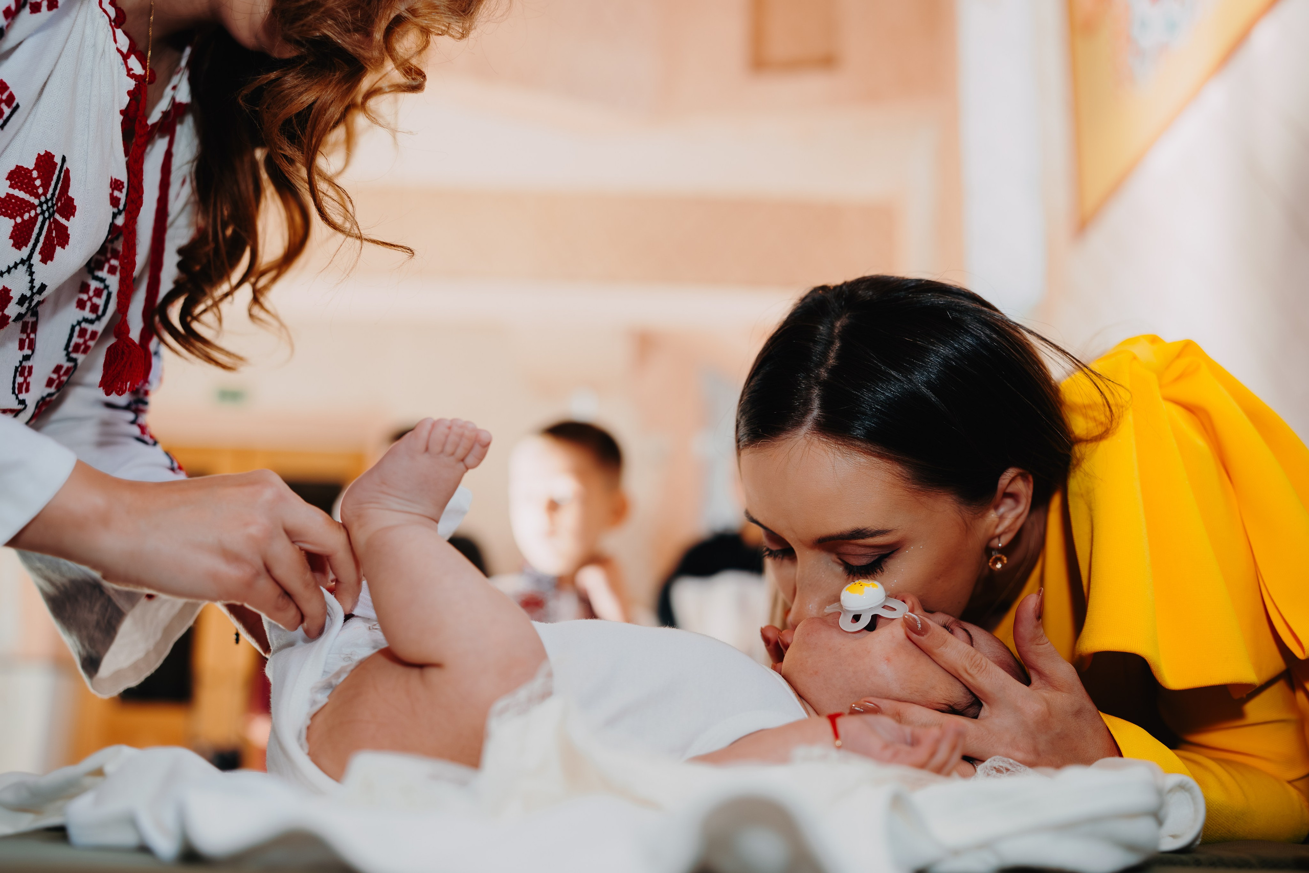 Moment tandru și uman în timpul botezului lui Arthur Mihail. Pe masa sfântă, mama îl sărută cu blândețe la cap pentru a-l calma, în timp ce nașa stă liniștită la picioarele lui. O poză autentică care captează emoția și conexiunea maternă chiar și în cadrul ritualului.