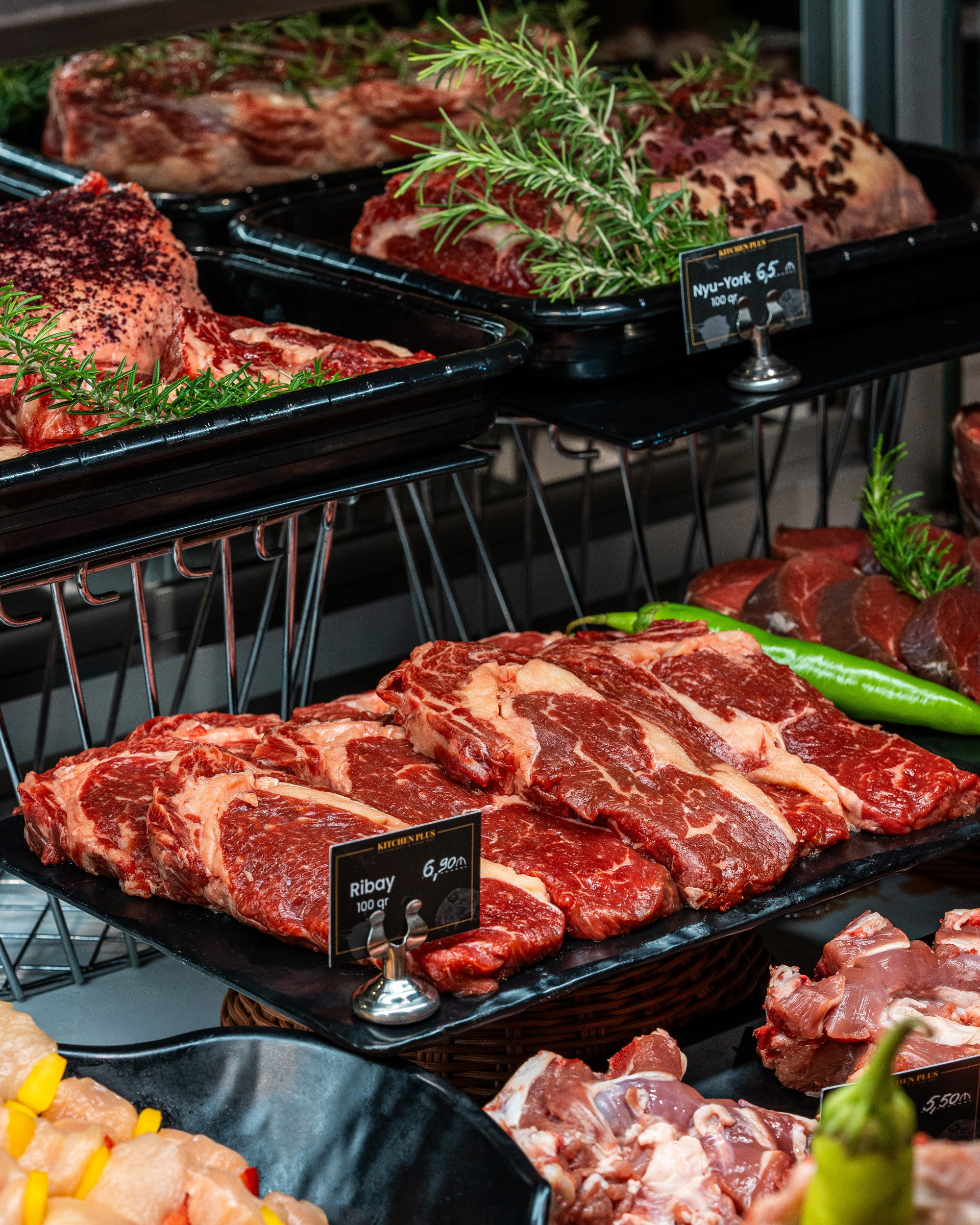 Qara tepsidə yığılmış bir neçə mərmərli Ribeye steyk dilimləri, üzərində qiymət etiketi, yaşıl bibər və rozmarin.