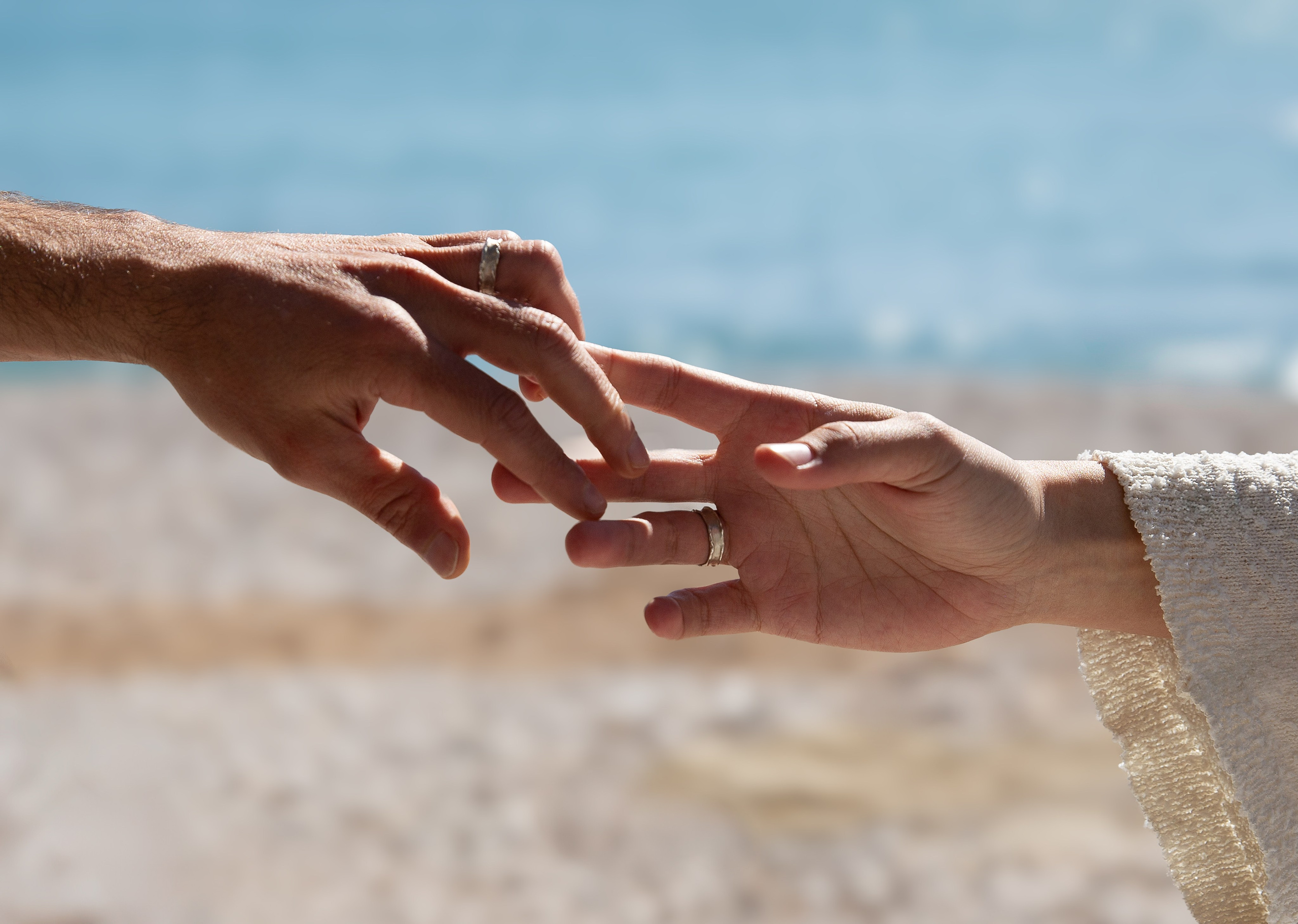 Close-up photo of a couple’s hands gently reaching for each other, both wearing wedding rings, with the soft beach sand and ocean waves in the background, symbolizing connection and love Valencia, Spain.