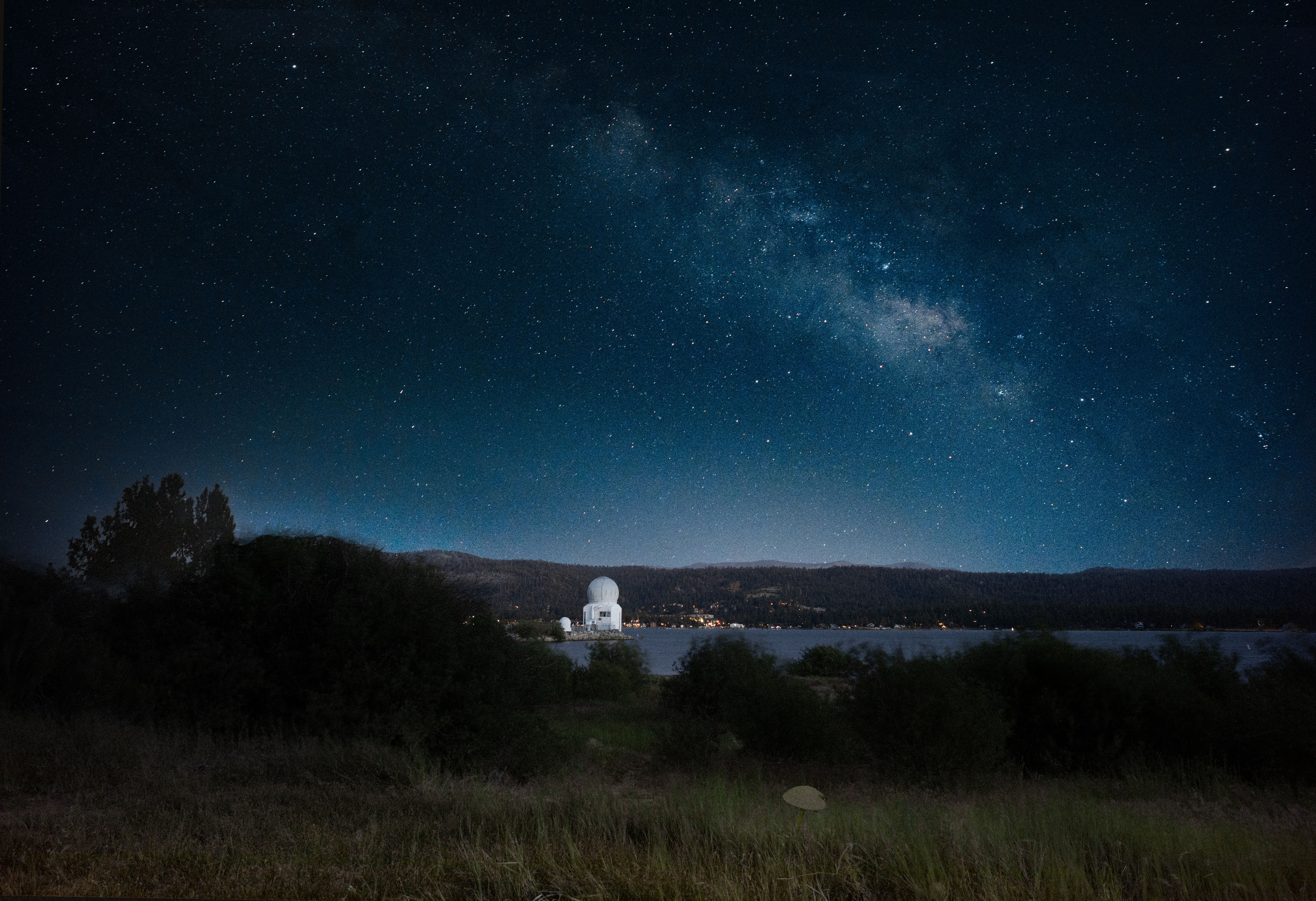From the quiet forested shore of Big Bear Lake, the white dome of the Big Bear Solar Observatory glows softly against a star-filled sky. The stillness of the lake and the brilliance of the stars create a perfect contrast of earth and cosmos. While I stood alone capturing this image, two coyotes slipped silently through the darkness, pausing only to glance at me before vanishing again into the night—reminders that even in solitude, we are never truly alone under the stars.