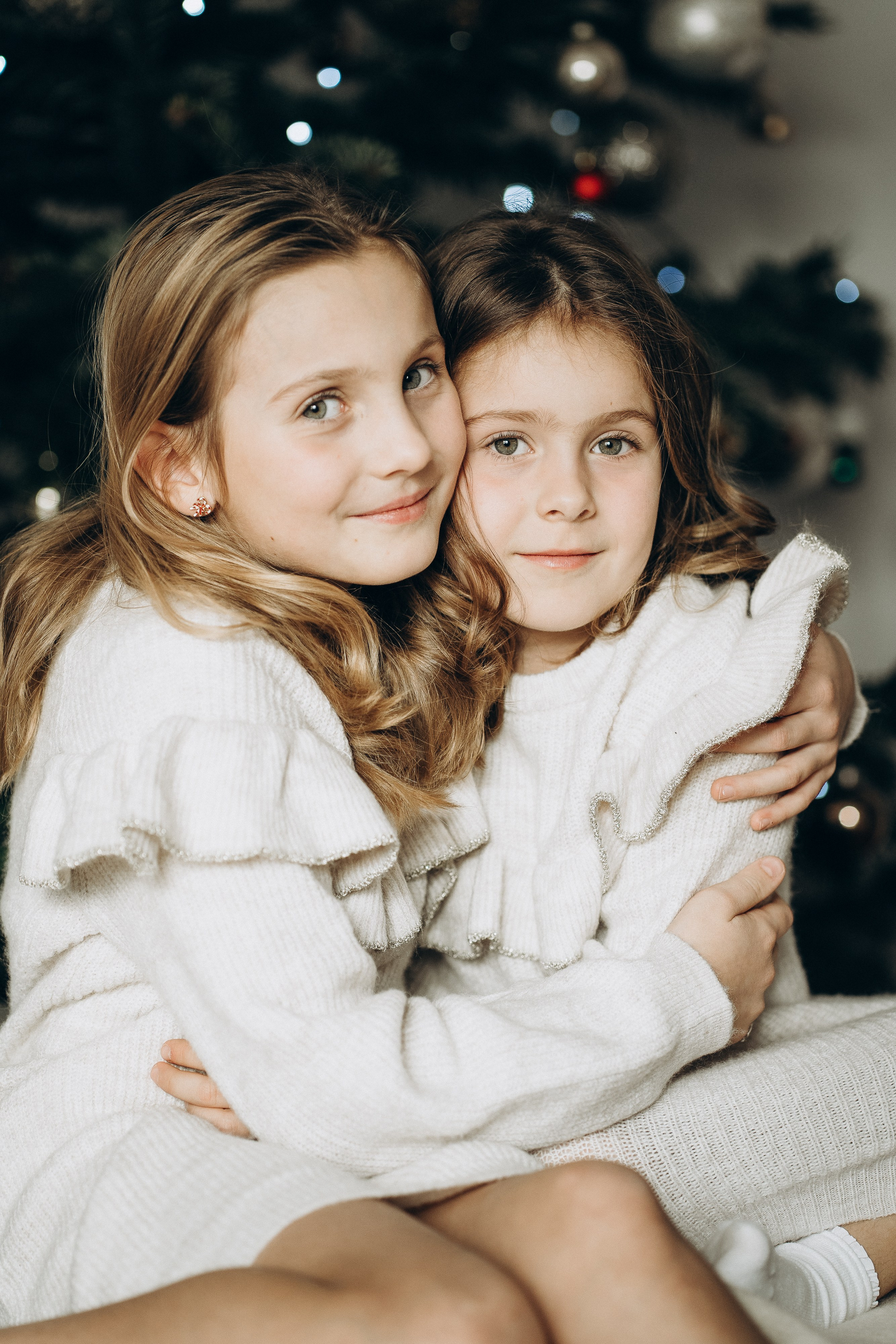 2 Schwester umarmen sich liebevoll vor dem Weihnachtsbaum.