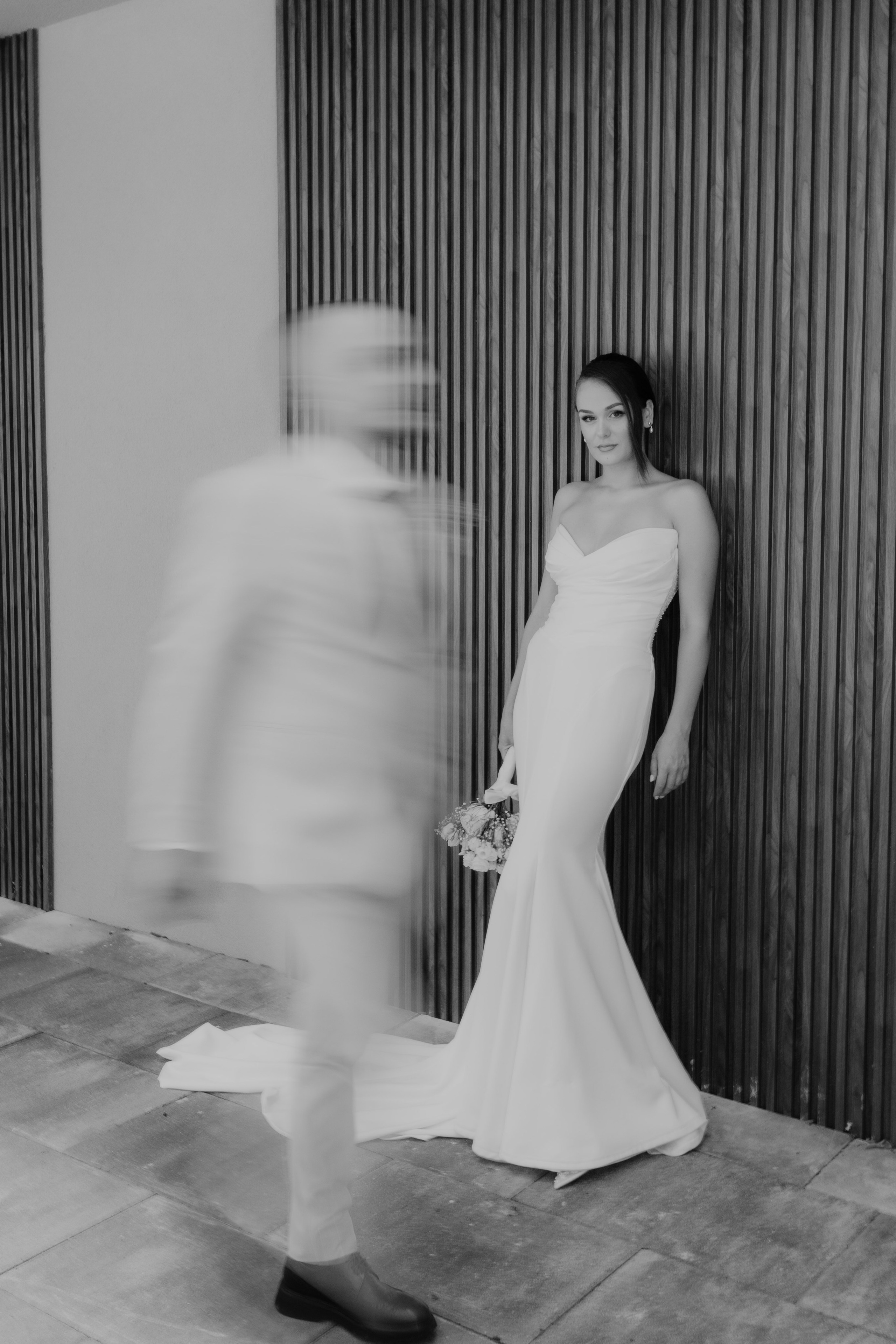 Elegant bridal portrait as the groom walks past in motion, creating an artistic and authentic wedding photography moment.