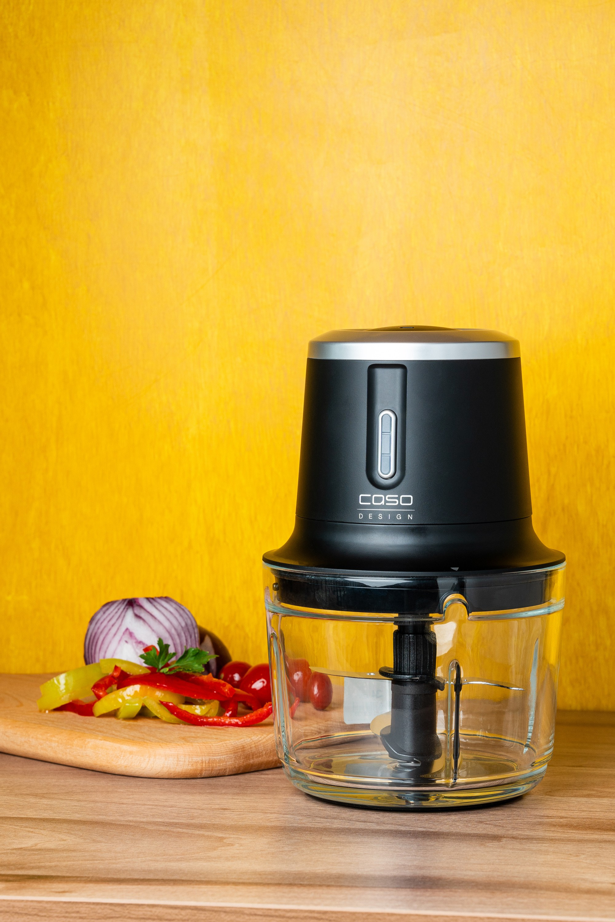 Caso Design elektrik doğrayıcı, sarı fon və kəsilmiş tərəvəzlərlə taxta səth üzərində təqdimat.