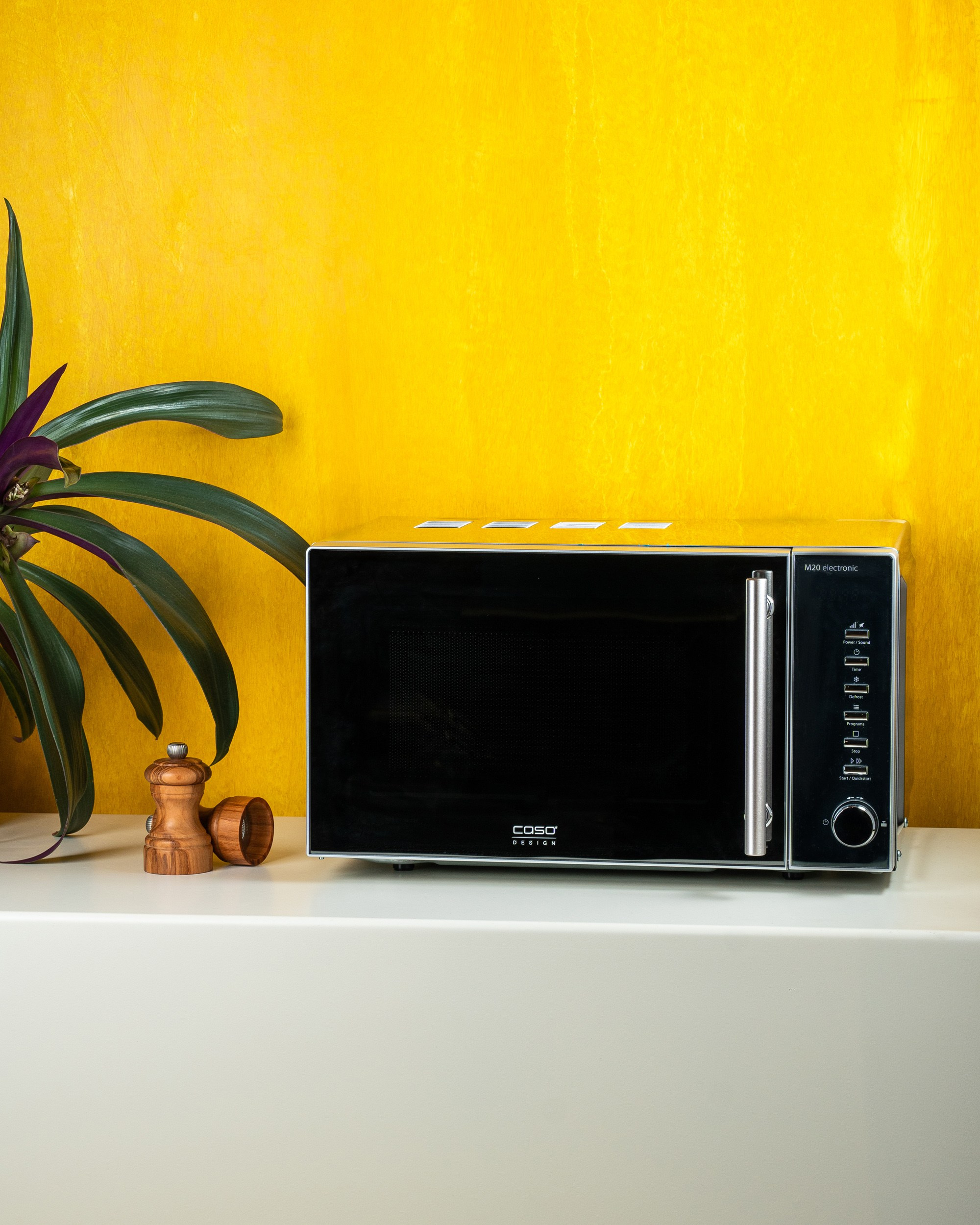 Caso Design mikrodalğalı soba, sarı fon və dekorativ bitki ilə ağ səth üzərində məhsul təqdimatı.