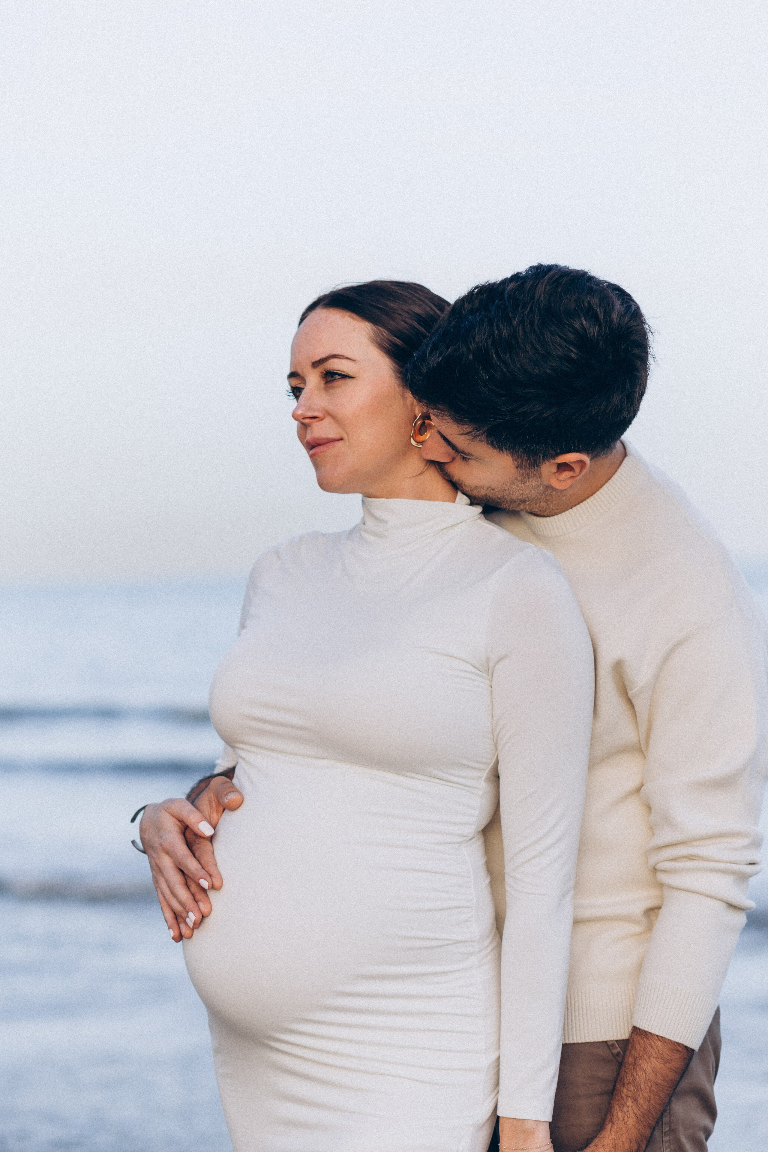 Retrato tierno junto al mar en Valencia, España — pareja abrazada mientras el compañero besa el cuello de la futura madre, ambos vestidos en tonos neutros durante una tarde tranquila en la costa.