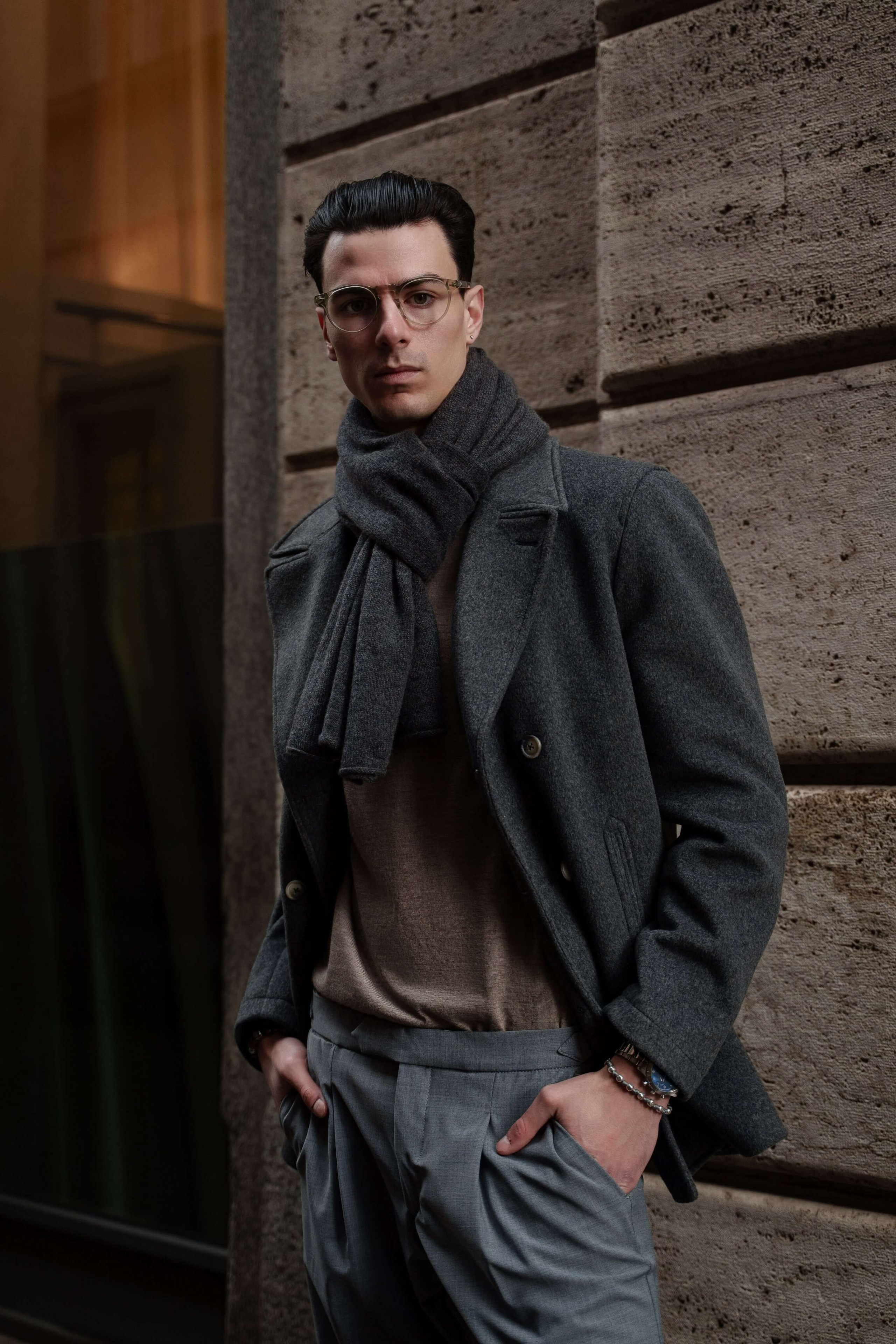 Stylish man in gray overcoat and scarf posing against stone wall in urban Milan
