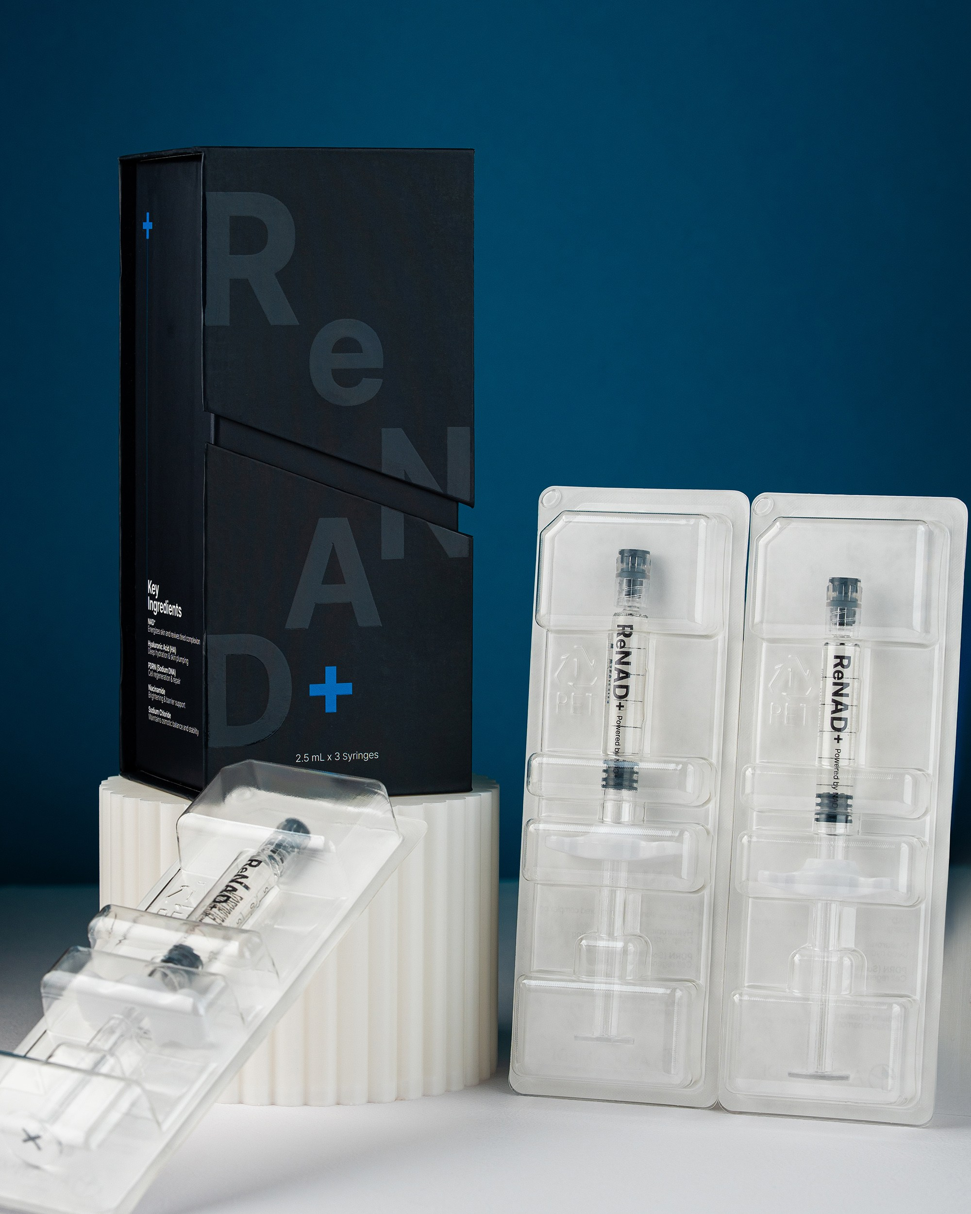 A professional shot of ReNAD+ skin booster featuring a sleek black box on a white pedestal, accompanied by three pre-filled syringes in clear blister packaging. Key ingredients like Hyaluronic Acid and PDRN are visible on the box.