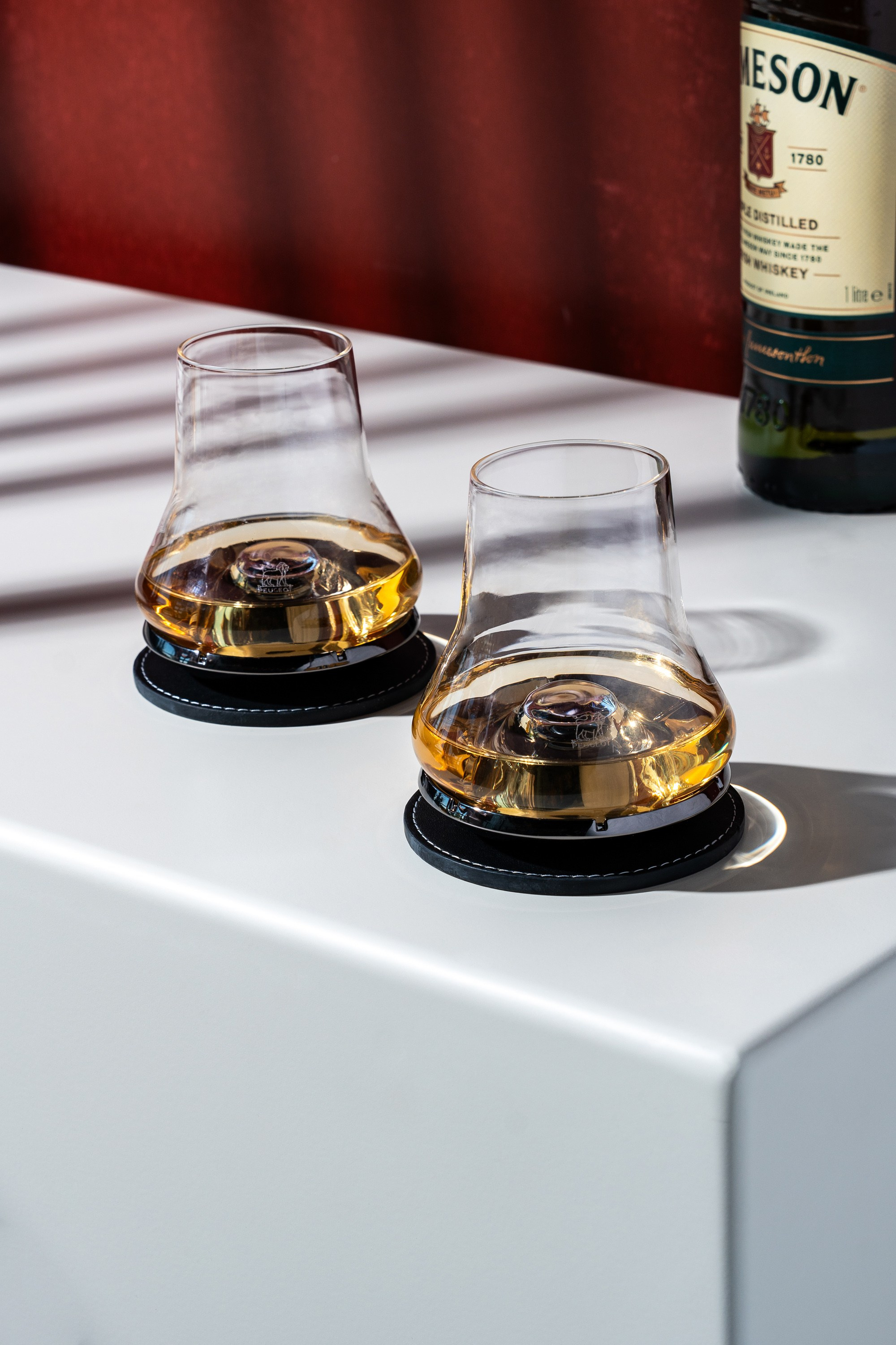 Ağ səth üzərində qara altlıqlarda dayanmış, viski ilə dolu iki "Glencairn" dequstasiya stəkanı, arxa planda Jameson şüşəsi.