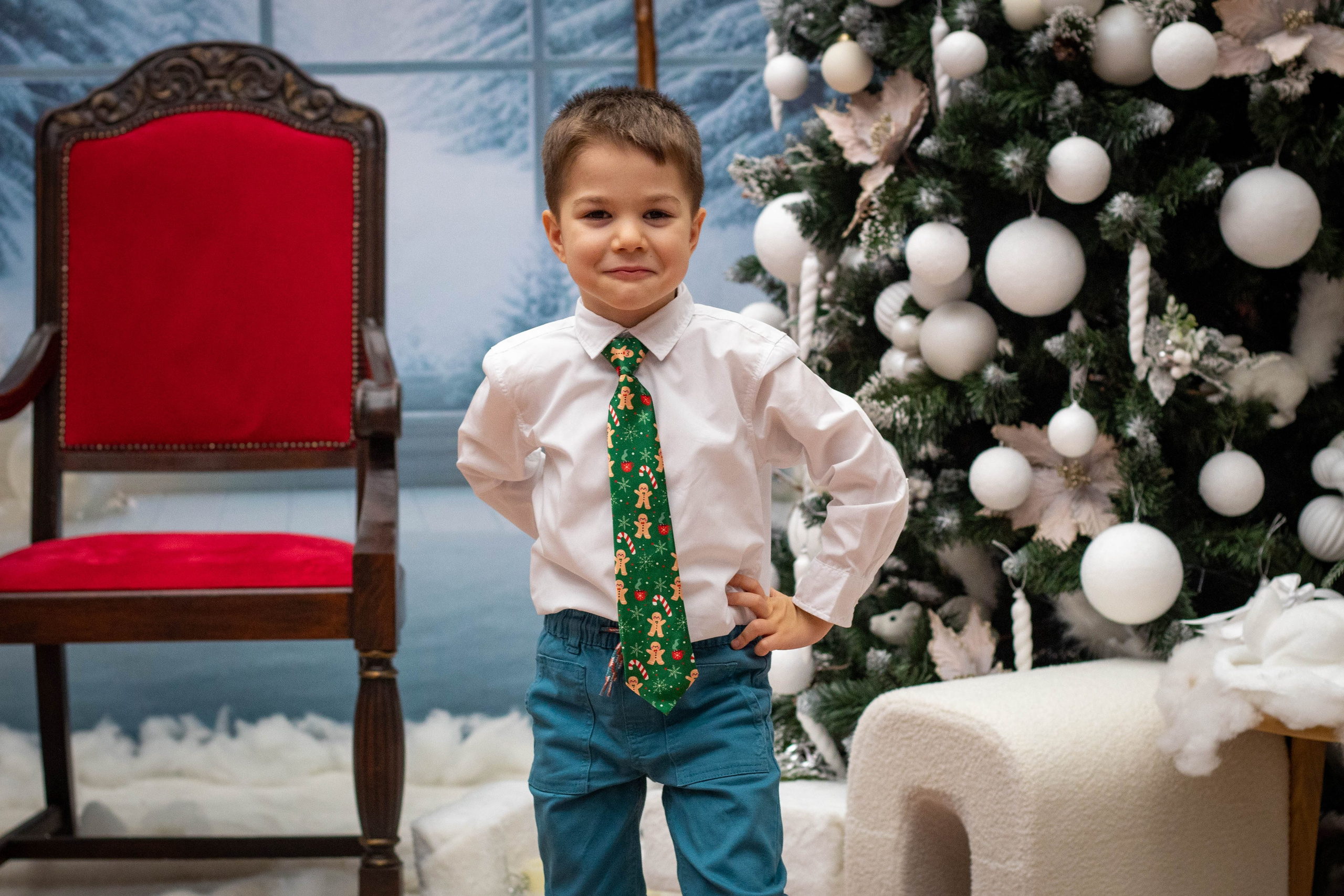 Young boy in festive clothes sitting on a decorative red chair in a holiday backdrop.