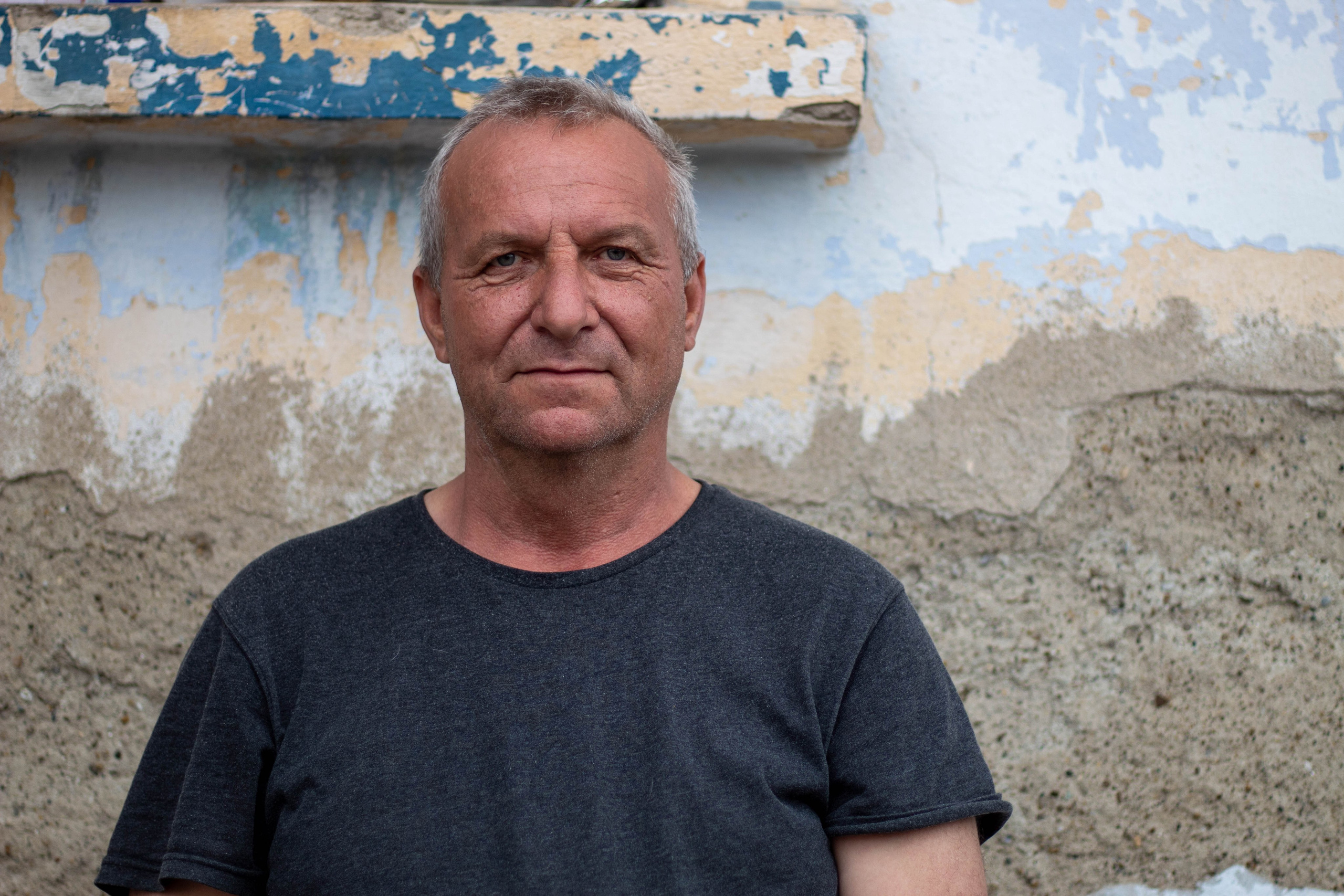 Portrait of a man standing outdoors with a plain background, wearing a grey shirt and relaxed posture.