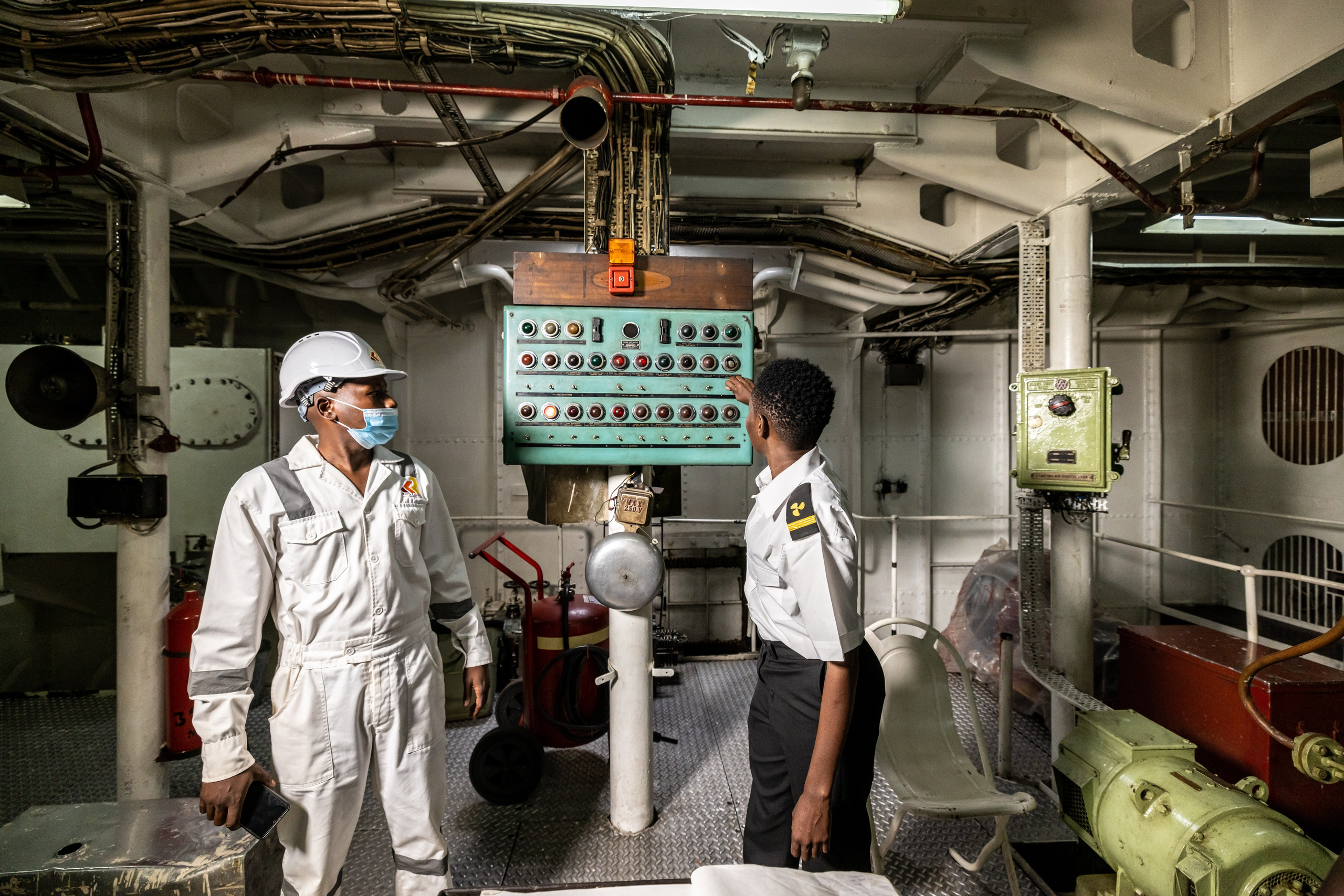 A wide shot of two engineers in the engine room of a ship, inspecting the engine. Documentary photography