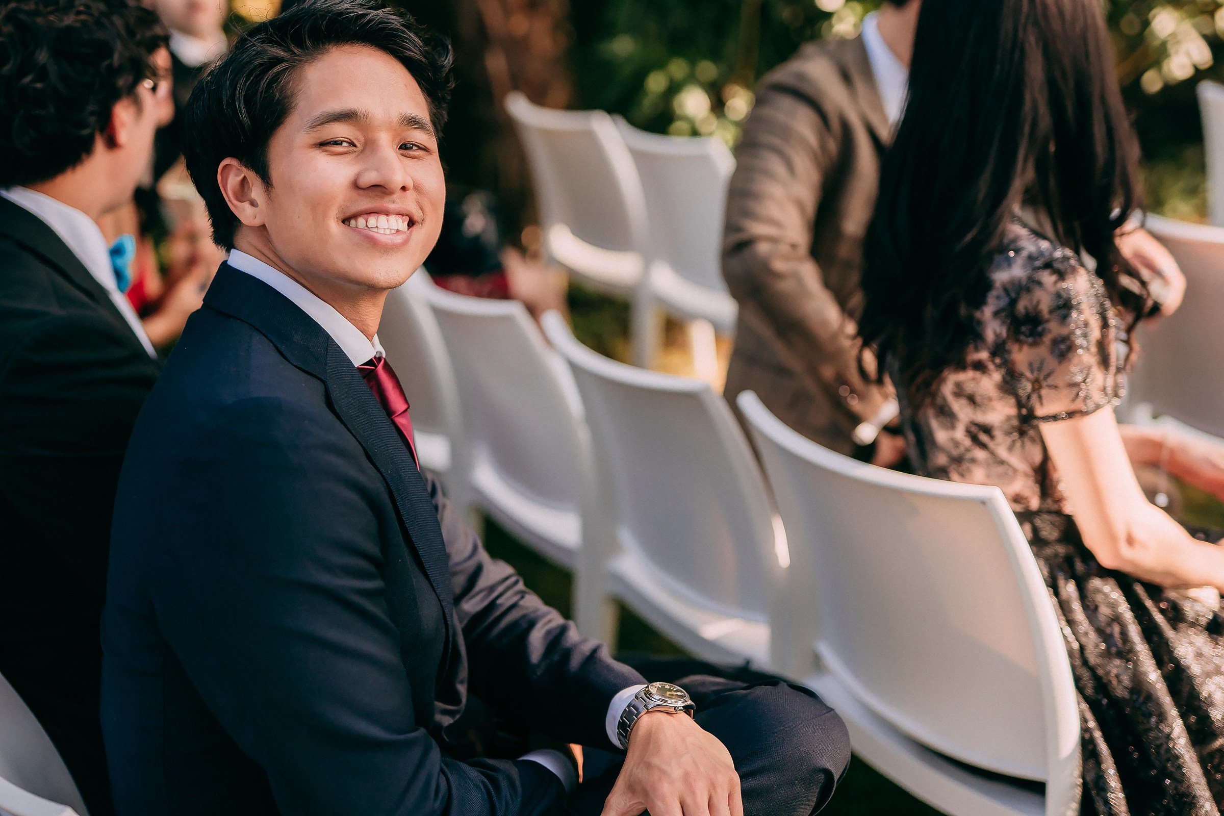 Young man in a navy suit smiles broadly while seated, enjoying the wedding ceremony.