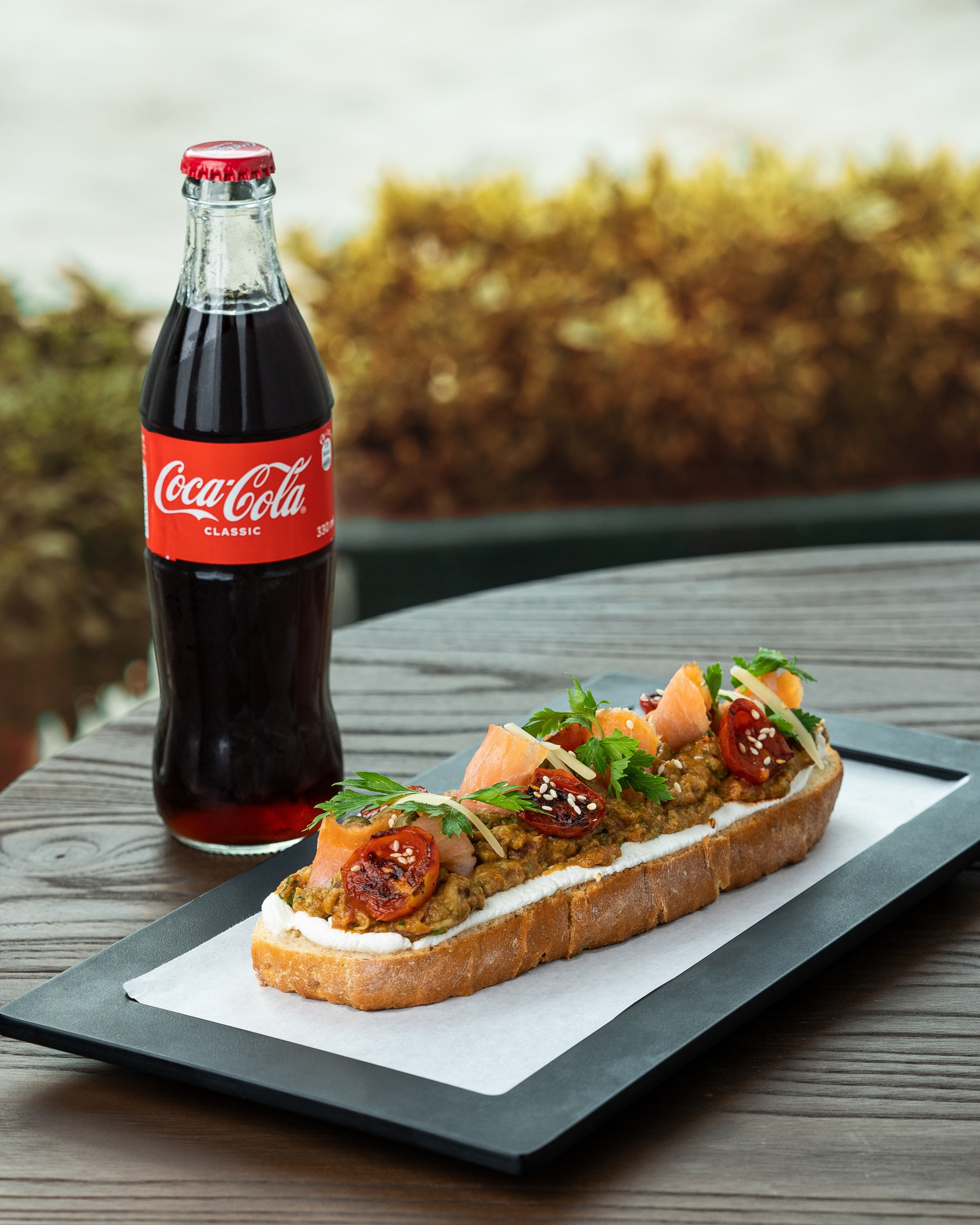 Qril tərəvəzlər, balıq və sousla bəzədilmiş açıq sendviç, yanında Coca-Cola şüşəsi.