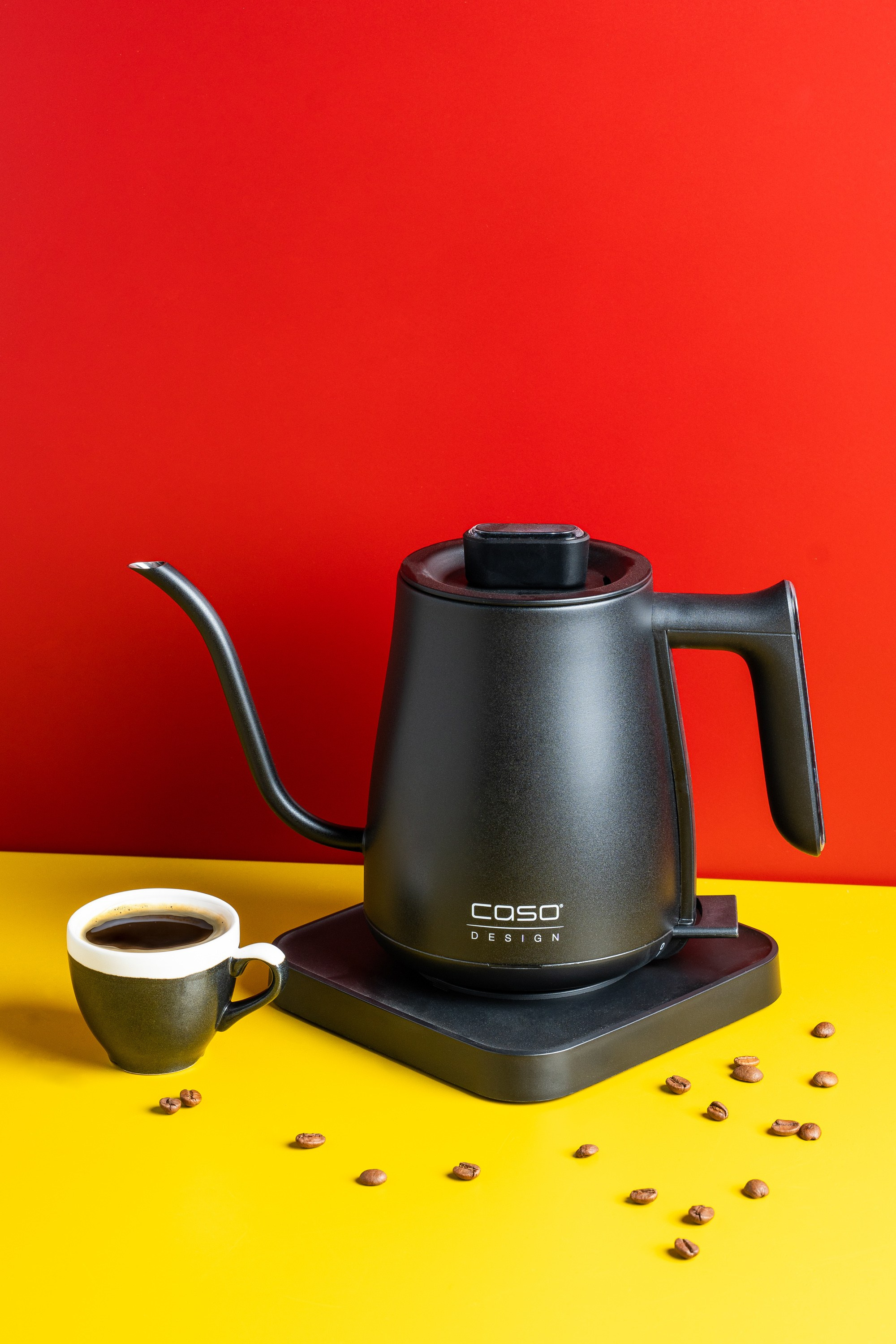 Caso Design elektrik qəhvə qızdırıcısı və qəhvə fincanı, qırmızı və sarı fon üzərində, qəhvə dənələri ilə birlikdə.
