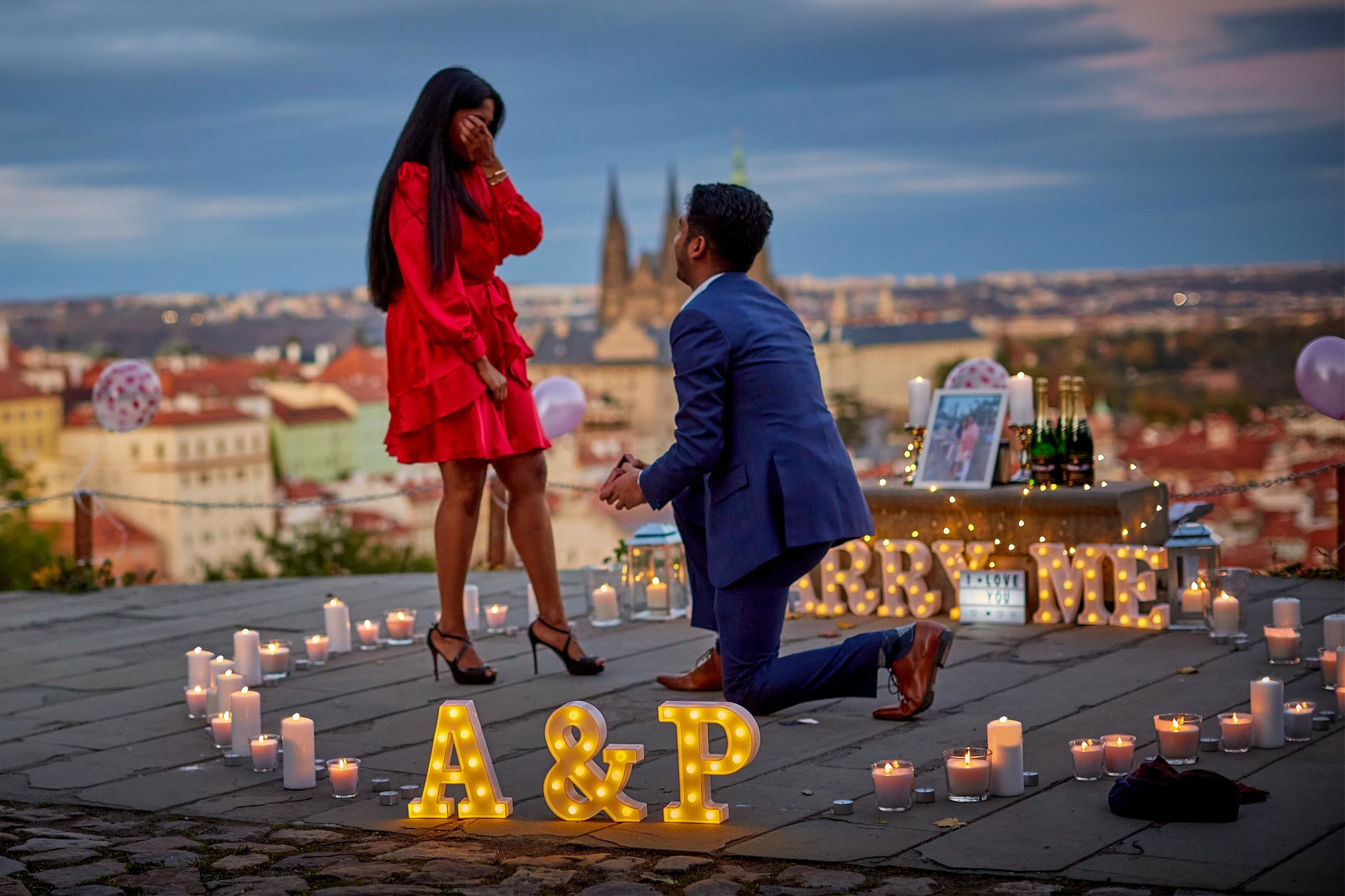 Woman in red dress wiping tears surrounded by candles and fairy lights at Prague proposal.