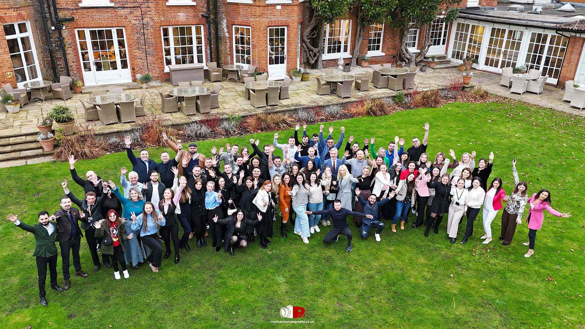 A large group of smiling corporate event attendees posing outside a charming brick building.