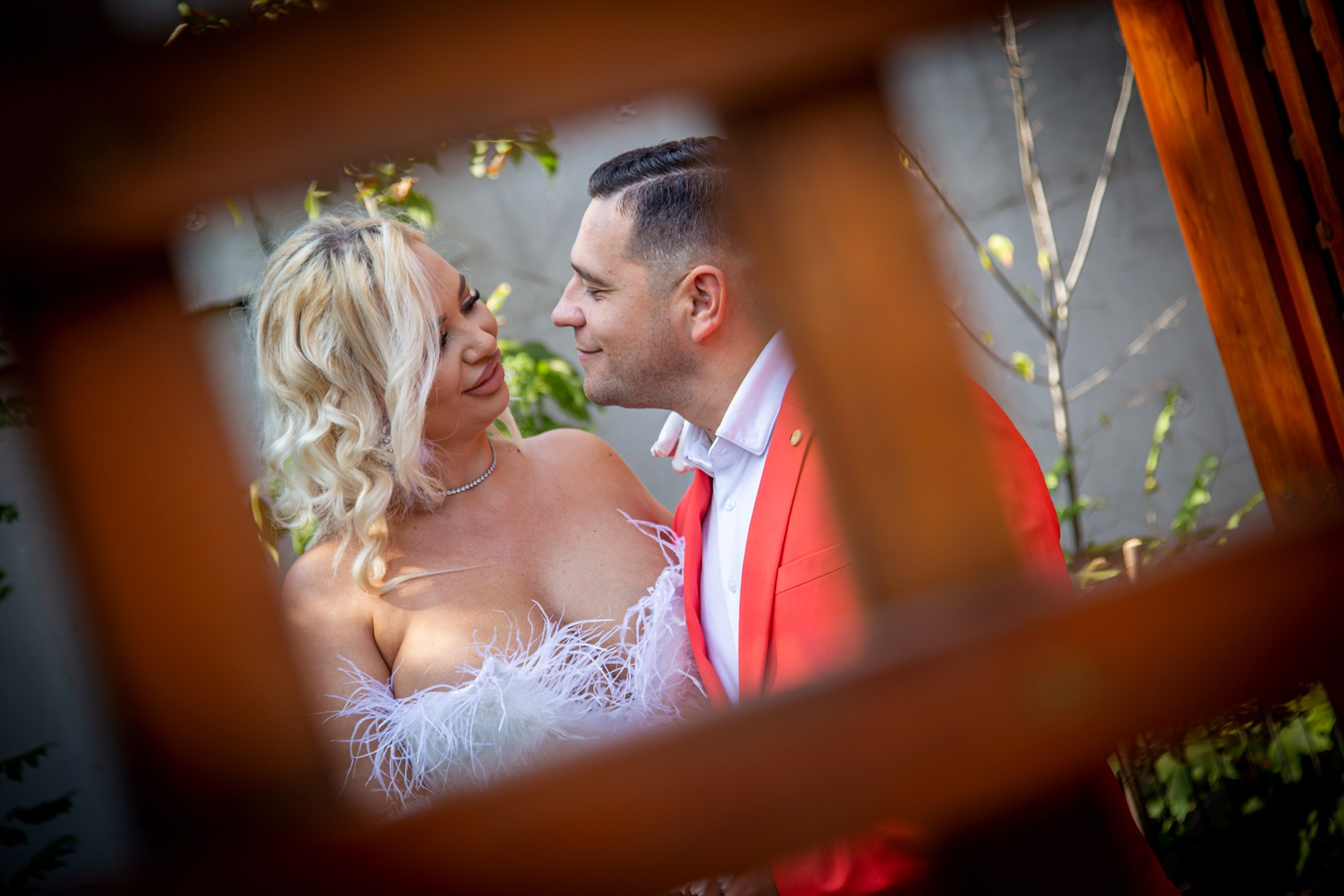 Cuplu de miri la ședința foto de la starea civila– mireasa în rochie albă cu pene și mirele în sacou roșu, poză romantică în aer liber