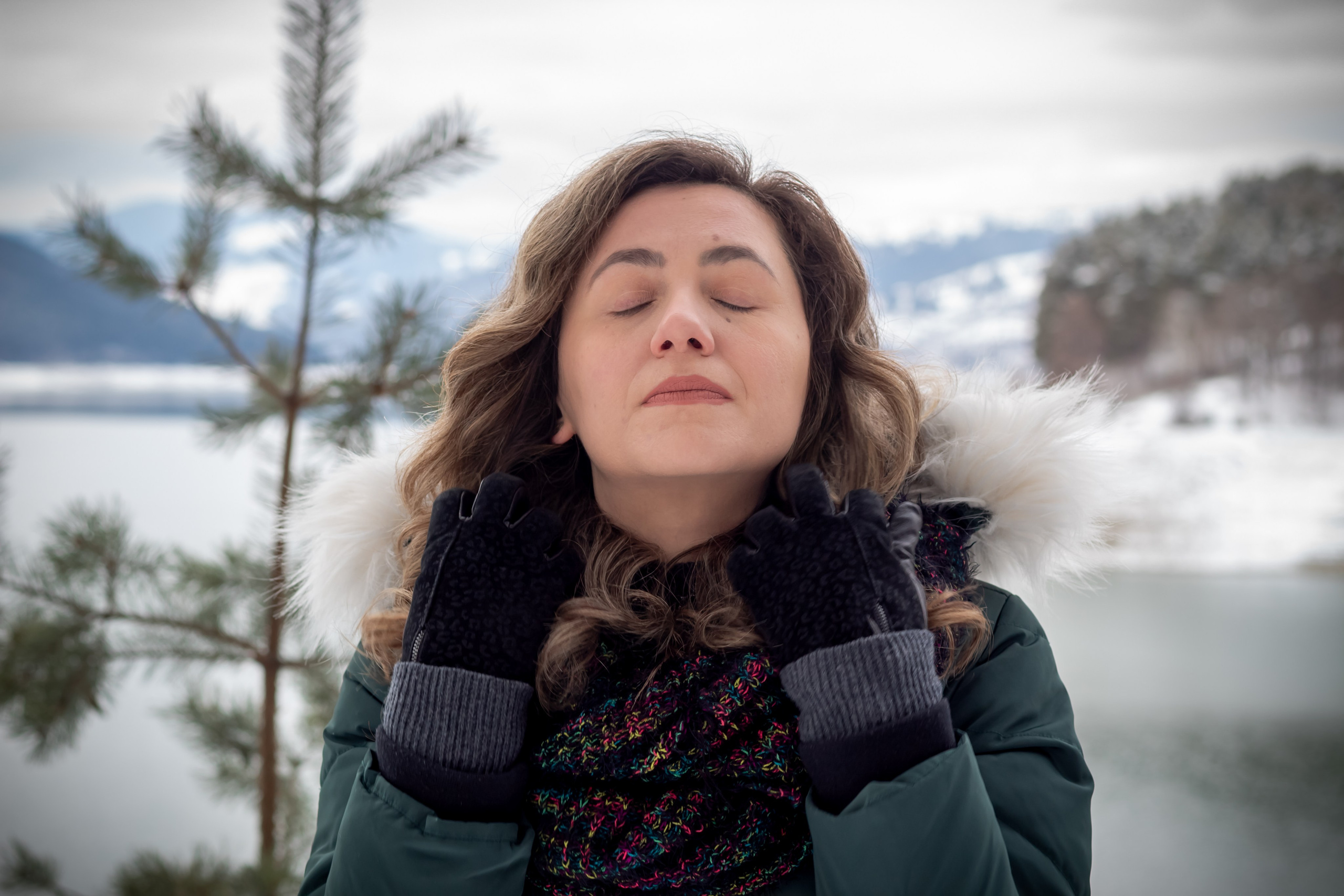 Woman with eyes closed standing near a lake in winter, wearing a scarf and coat, enjoying the crisp air.